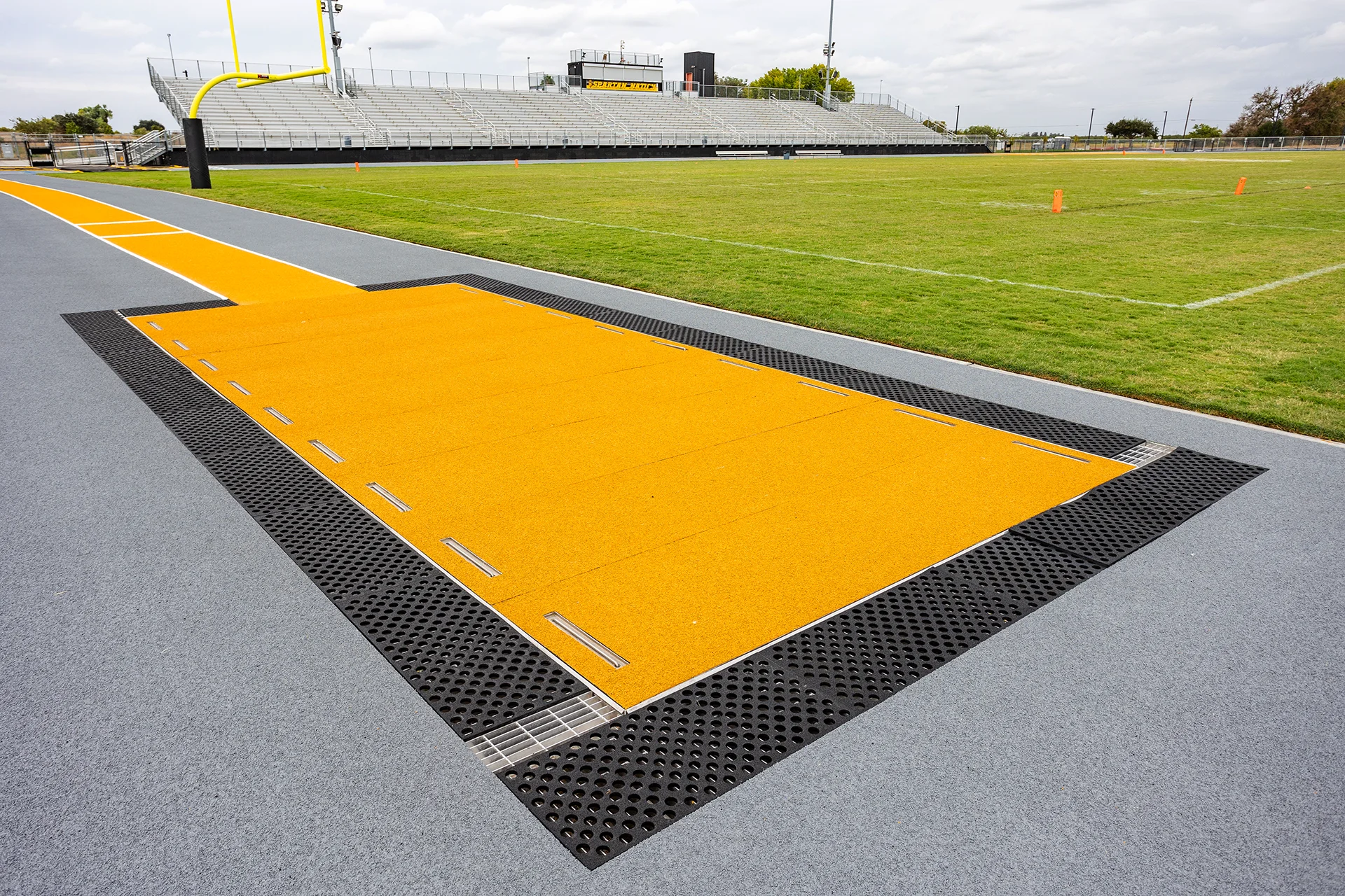 Long jump runway and takeoff area at Lathrop High School track and field complex with the football field, goalpost, and stadium bleachers in the background.