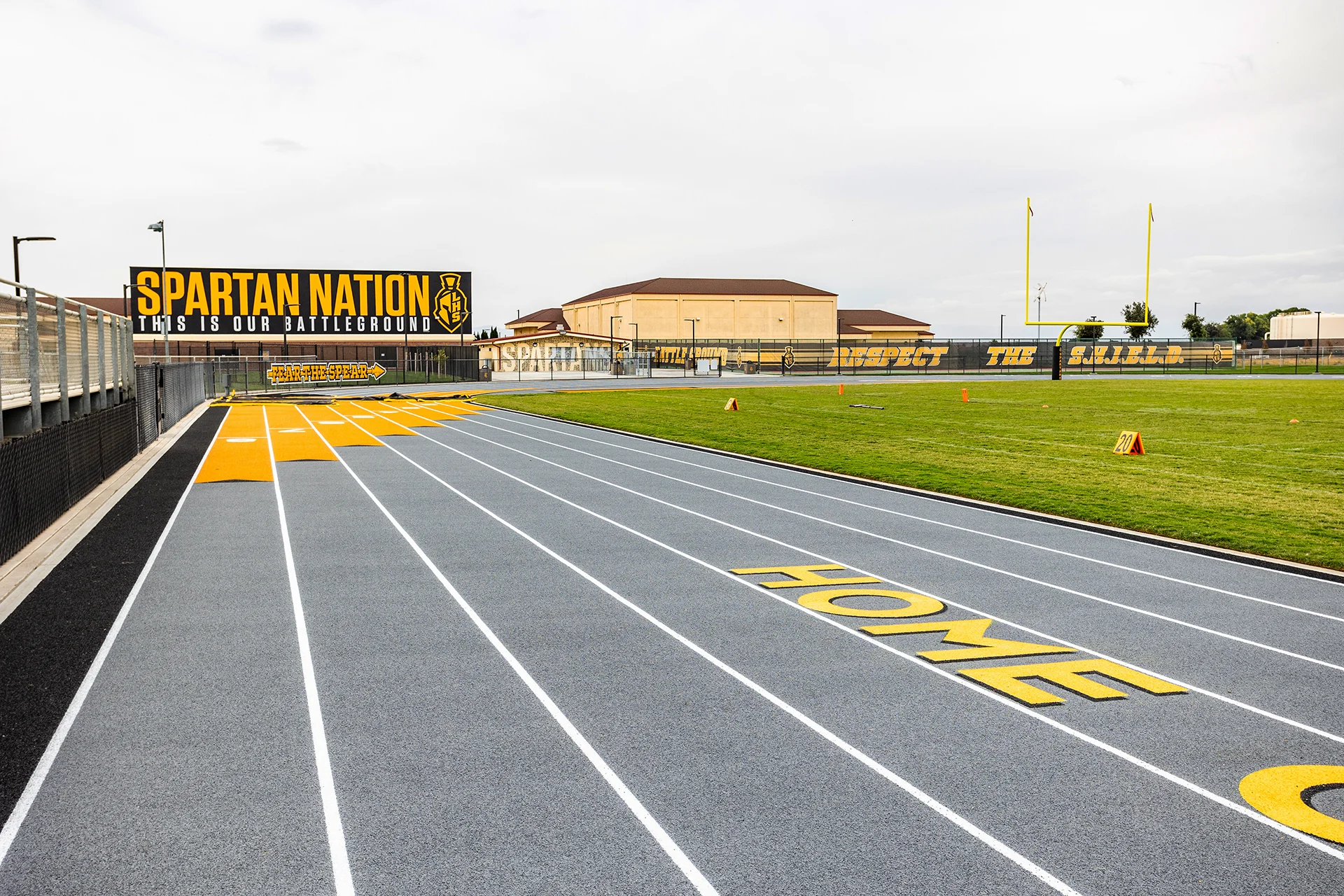 Lathrop High School running track beside the football field featuring Spartan branding, goalposts, and stadium signage in black and gold school colors.