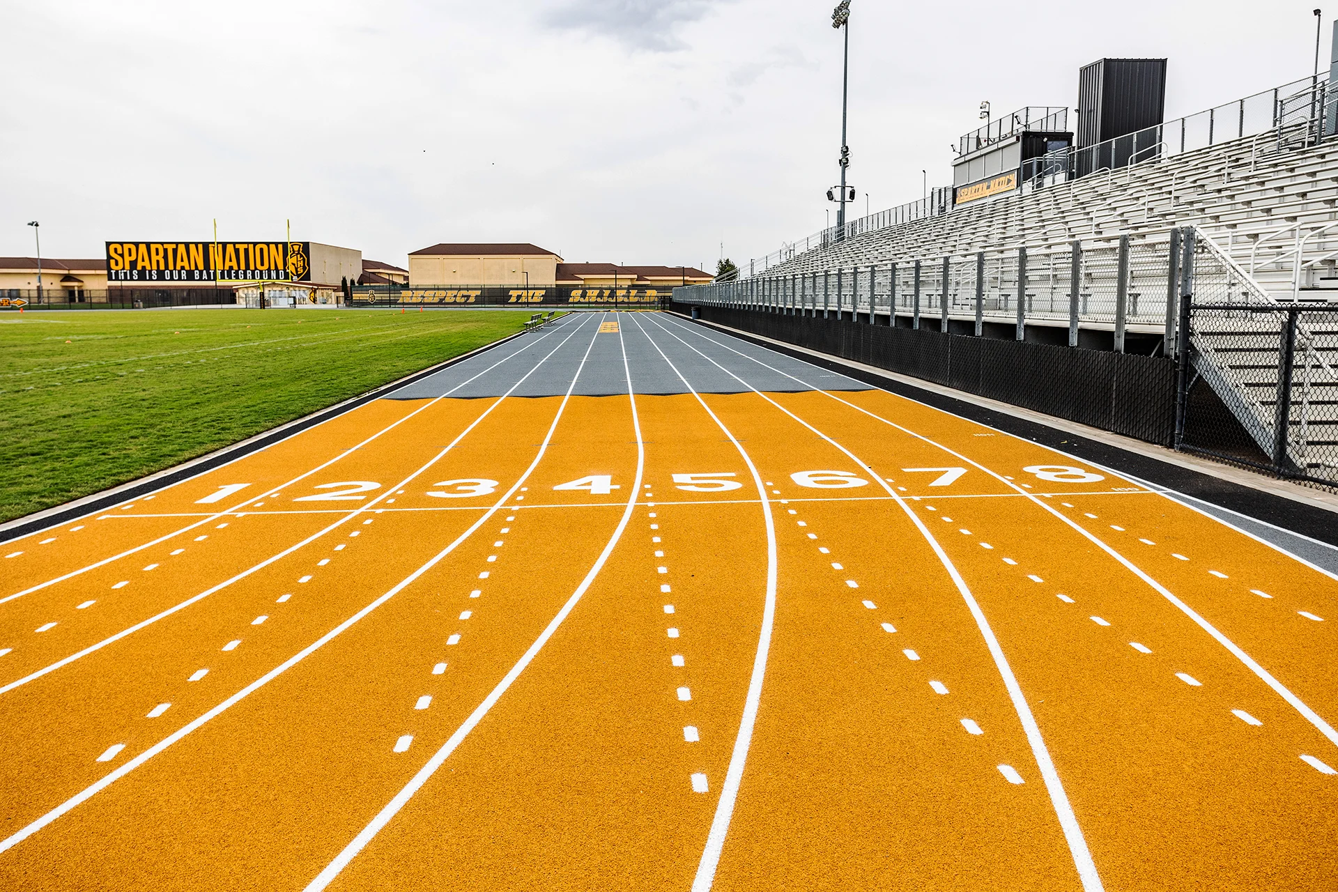 Lathrop High School running track featuring numbered starting lanes, stadium bleachers, football field, and Spartan-branded signage in black and gold school colors.