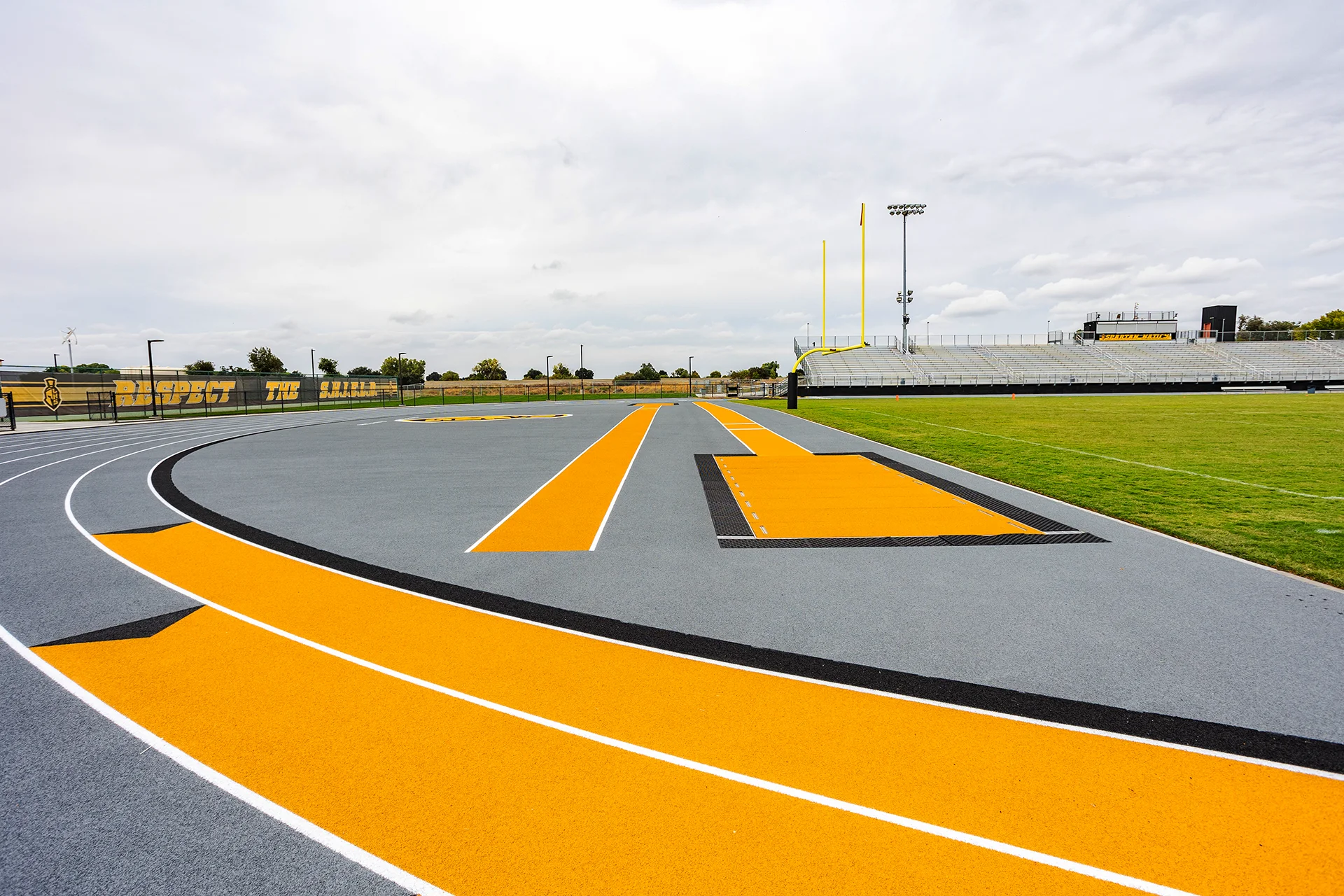 Lathrop High School track and field facility featuring a long jump runway, football field, goalposts, and stadium bleachers with black and gold school colors.