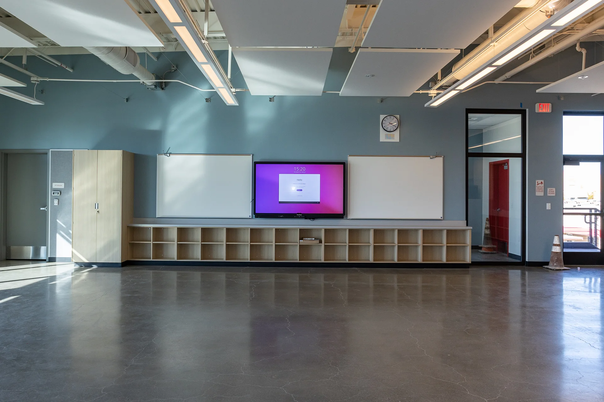 Expansive educational seminar room highlighting reflective sealed concrete floors, a central interactive smart monitor display mounted on a blue accent wall, custom low-profile open shelving cubbies, and suspended rectangular ceiling clouds.