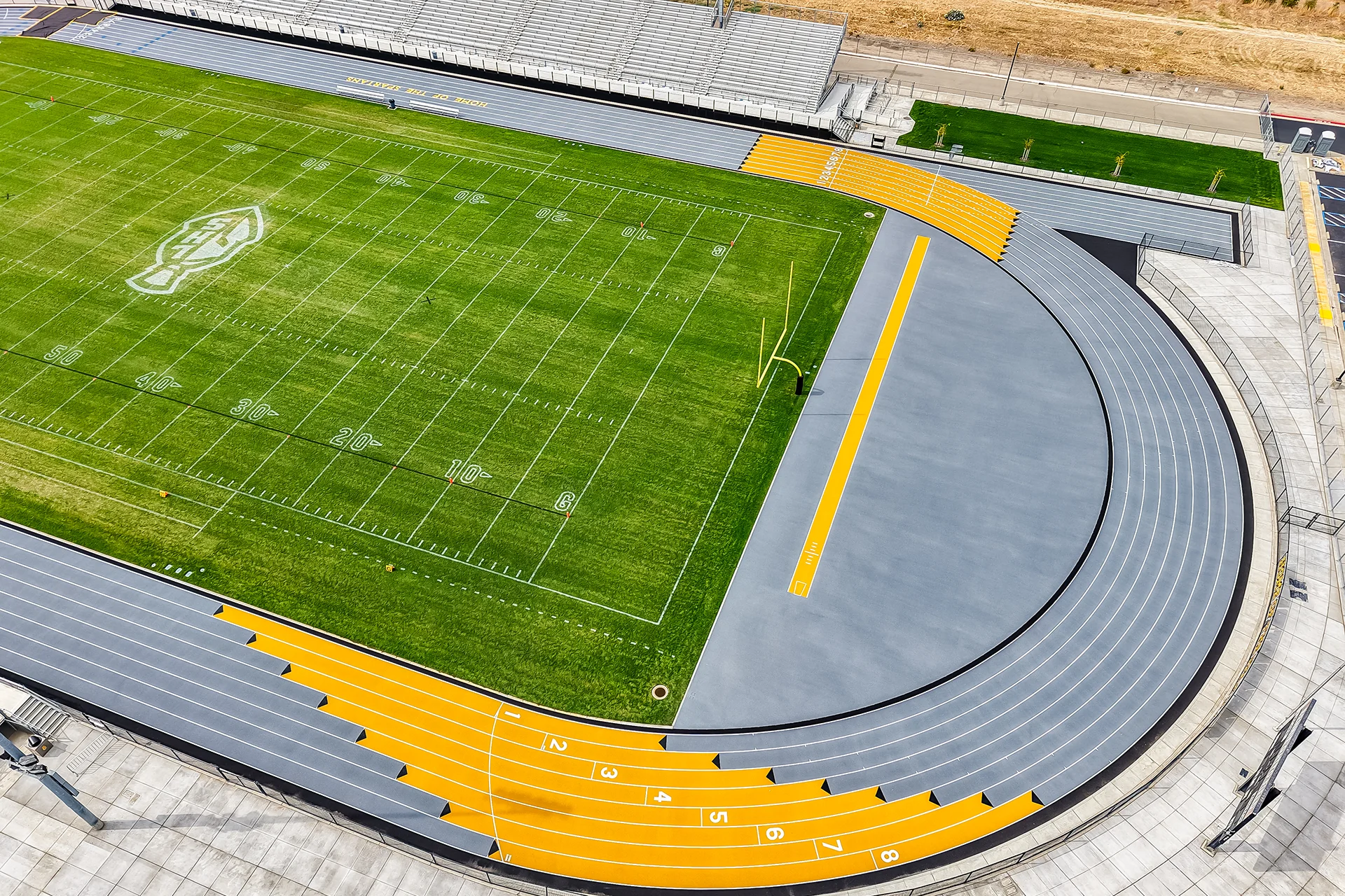 A low aerial photograph showing a grey running track curve and football field, with trees in the far distance under a bright, cloudy sky.