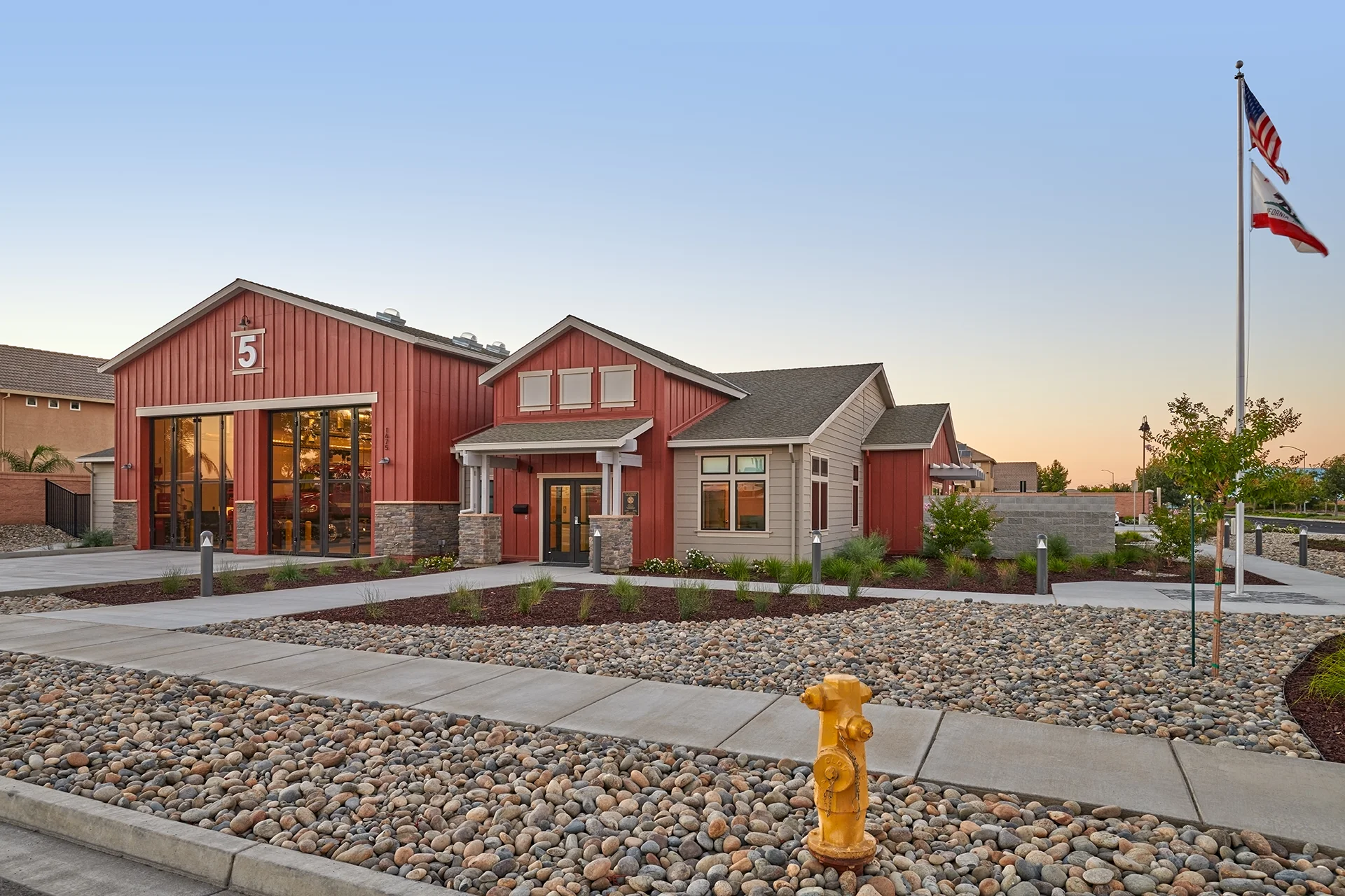 Landscape profile of the civic construction project at sunset, showcasing a yellow iron fire hydrant in the foreground, river rock groundcover beds, concrete public sidewalks, and the illuminated building exterior.