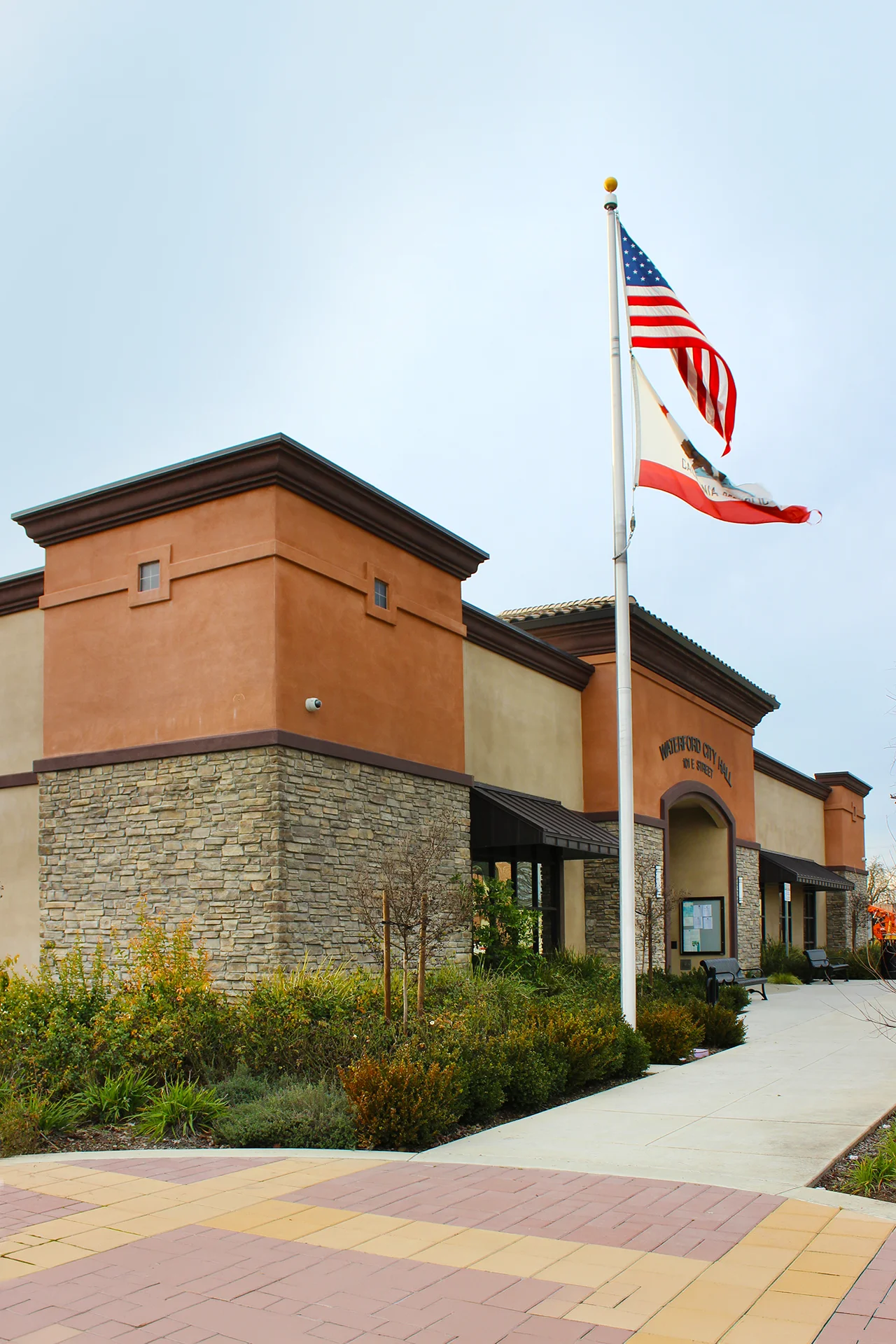 Daytime low-angle architectural perspective of the municipal building's exterior corner, showing beige stucco and orange accents, split-face concrete stone veneer on the pillars, an aluminum flagpole flying the United States and California Republic flags, and landscaped shrubbery.
