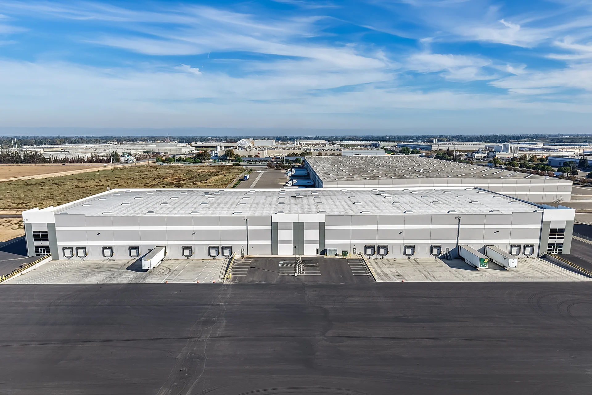 High aerial panorama capturing an expansive industrial park development, featuring white single-ply commercial roof assemblies with integrated smoke hatches, deep concrete dock pits, asphalt drives with tire marking signals, and surrounding industrial manufacturing zones.