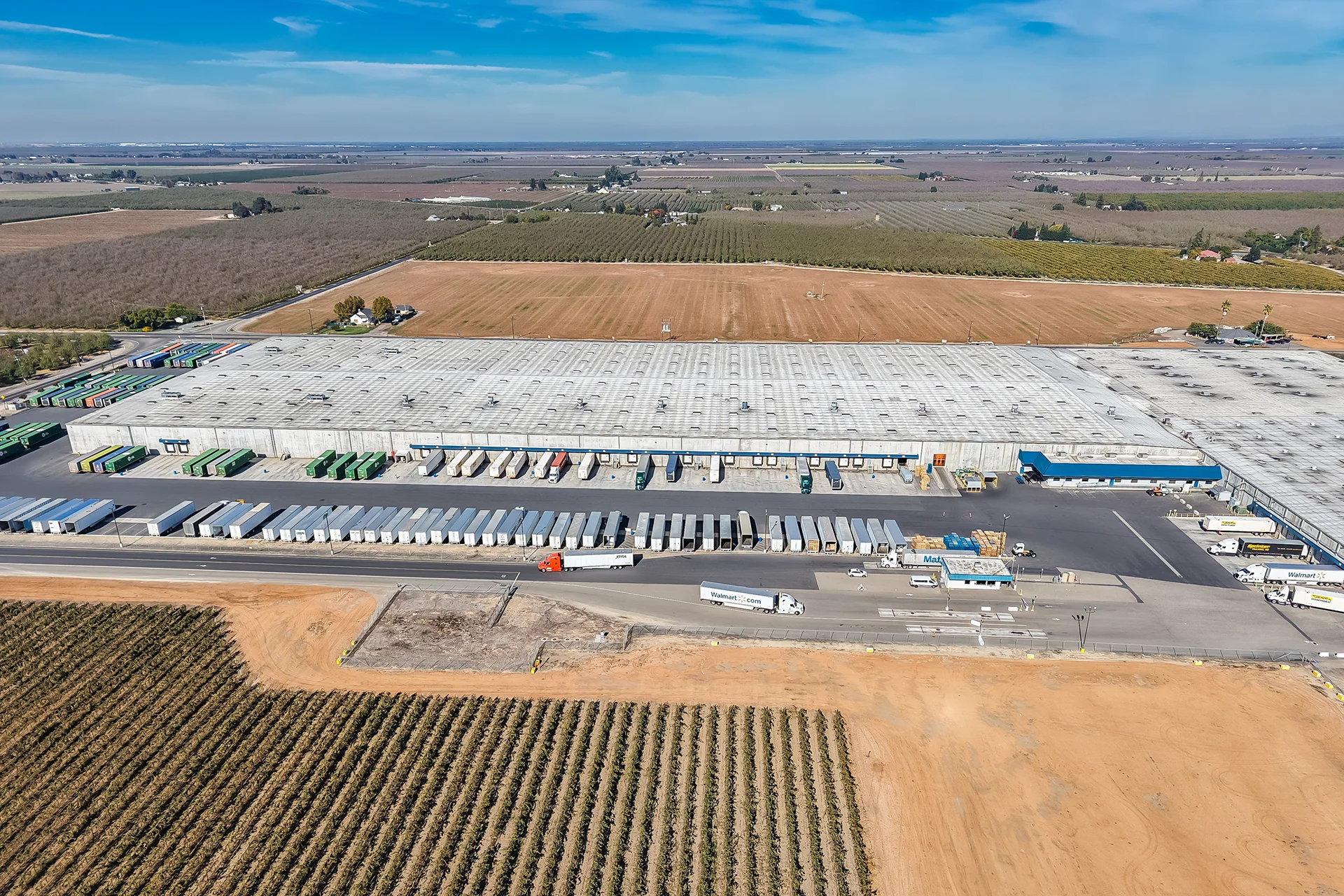 Comprehensive high-altitude drone shot of a major agricultural processing and distribution facility, showcasing a massive footprint warehouse with dozens of distribution docks, a central truck scale entry, active fleet trucks on asphalt thoroughfares, and cultivated vineyard rows in the foreground.