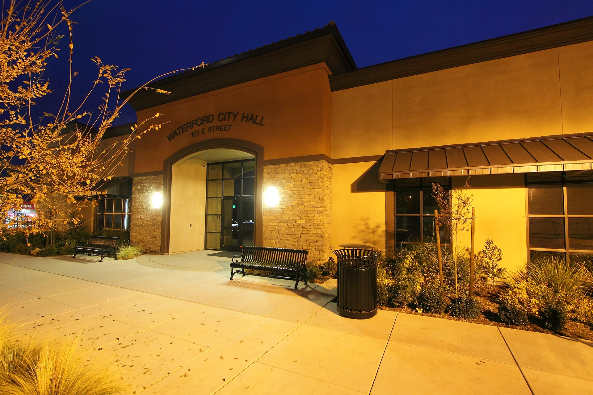 Nighttime architectural shot of the building's central entryway, highlighting a large stucco archway displaying Waterford City Hall 101 E Street, flanked by stacked stone veneer walls and illuminated by bright outdoor wall sconces over black iron park benches.