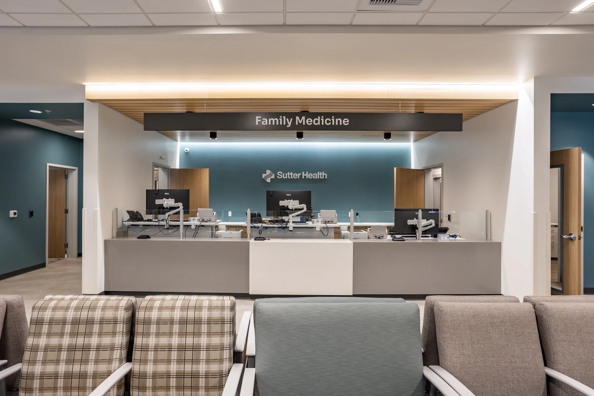 Direct eye-level view of an institutional medical registration desk equipped with triple computer monitors on articulating arms, built-in protective glass screens, solid surface white countertops, light oak accents, and a suspended linear metal and wood canopy with overhead spotlights.