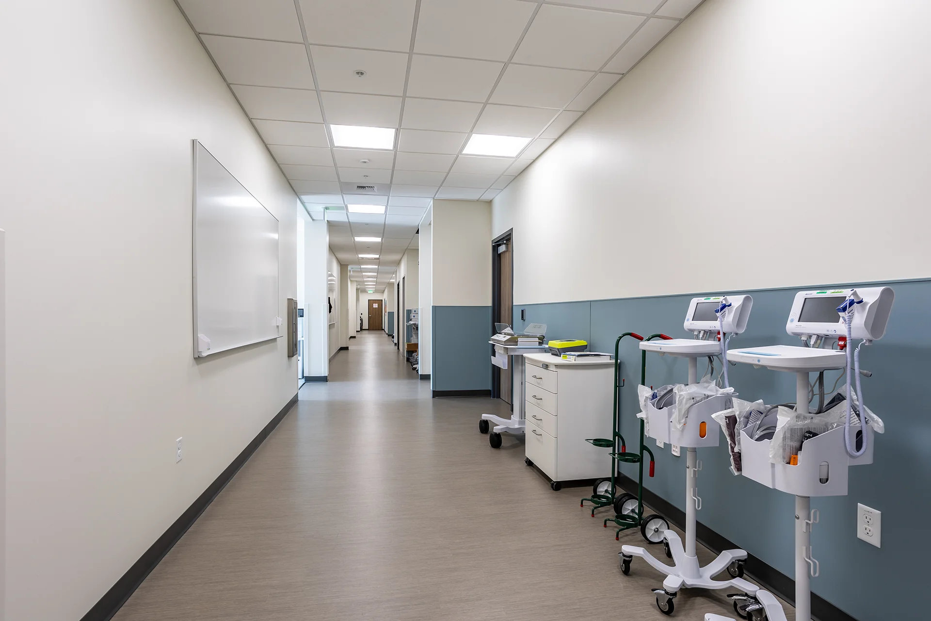 Expansive medical clinic hallway featuring durable vinyl plank flooring, a wall-mounted dry erase presentation board, teal vinyl wall protection panels, and multiple rolling Welch Allyn vital signs patient monitors plugged into commercial wall outlets.
