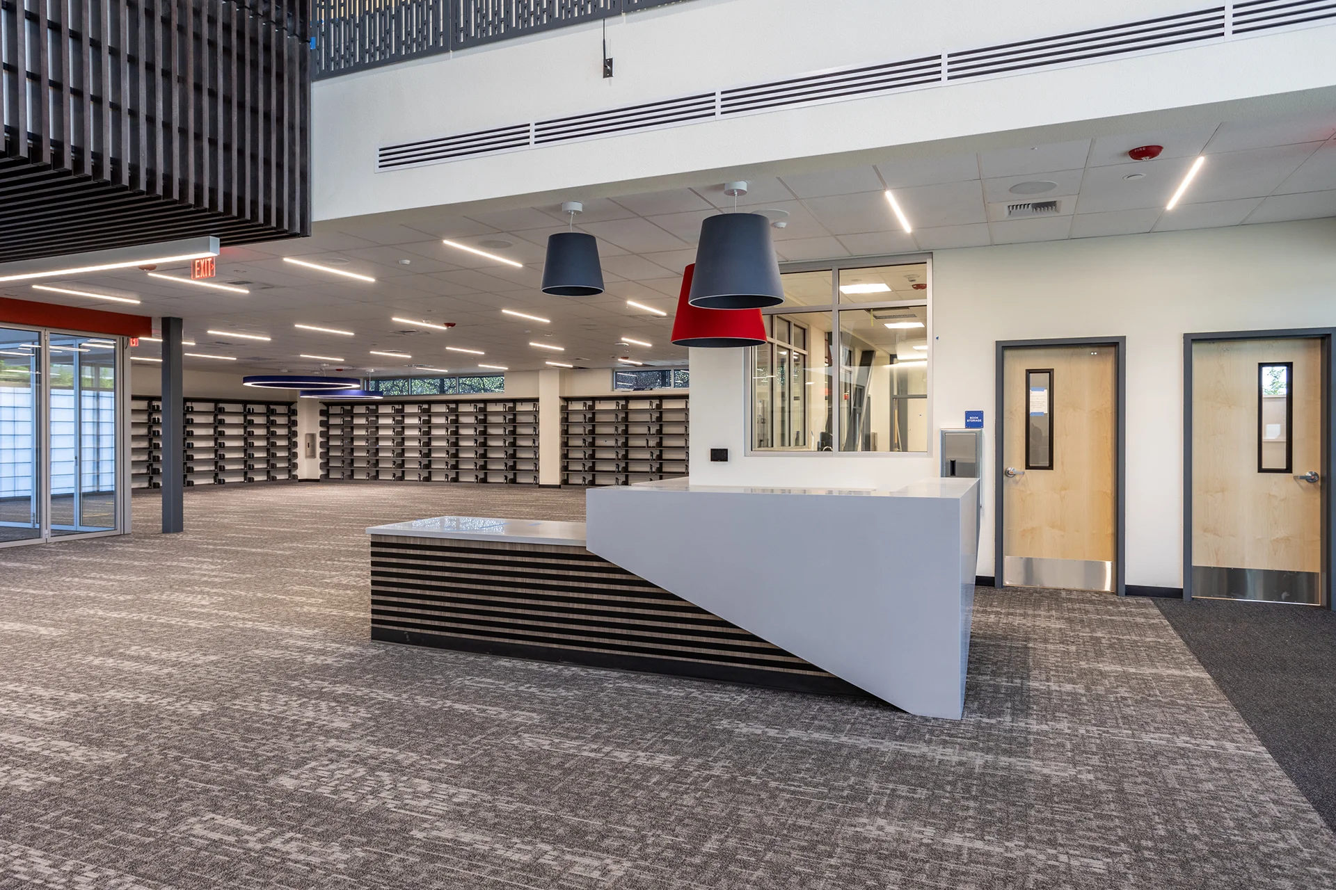 Front view of a geometric custom reception desk with laminate panel accents, modern oversized pendant light fixtures, wood entry doors with vision panels, and expansive perimeter wall shelving arrays inside an institutional school library.
