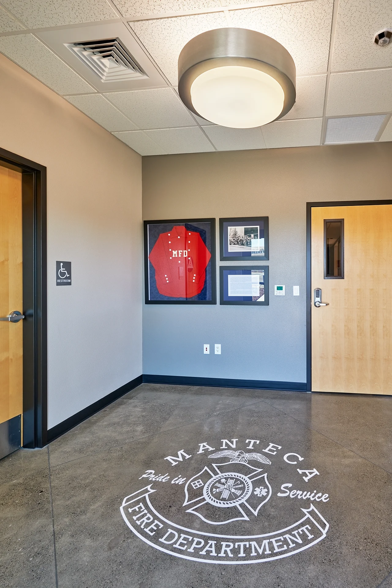 Main lobby entry alcove featuring the white engraved Manteca Fire Department logo on a polished concrete floor, custom-framed station memorabilia including a historic red uniform shirt, and access control wooden office doors.