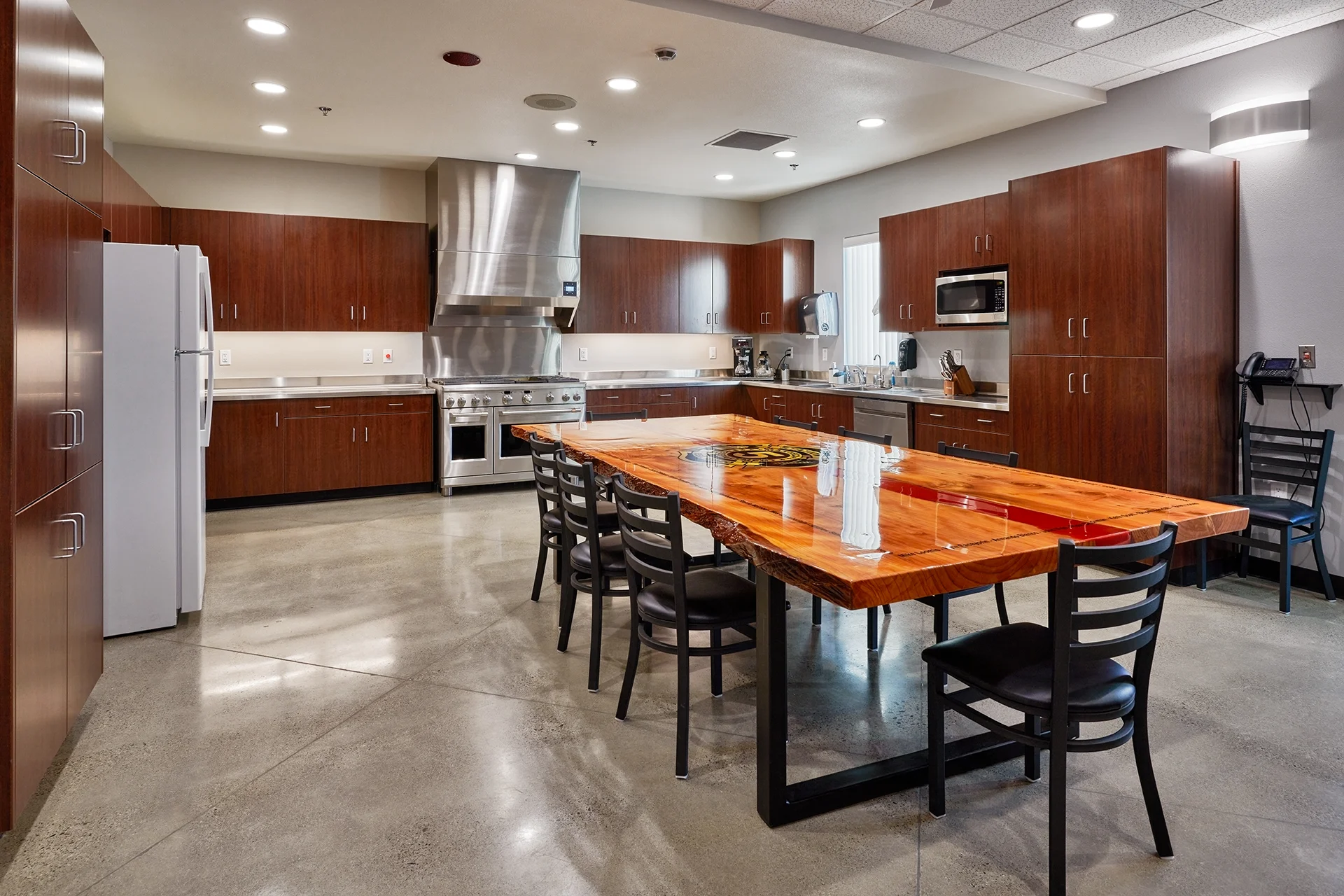 Commercial-grade kitchen facility inside Manteca Fire Station No. 5 featuring dark wood cabinets, stainless steel countertops, an industrial dual-oven range, and a polished live-edge wood dining table with built-in branding and black metal legs.