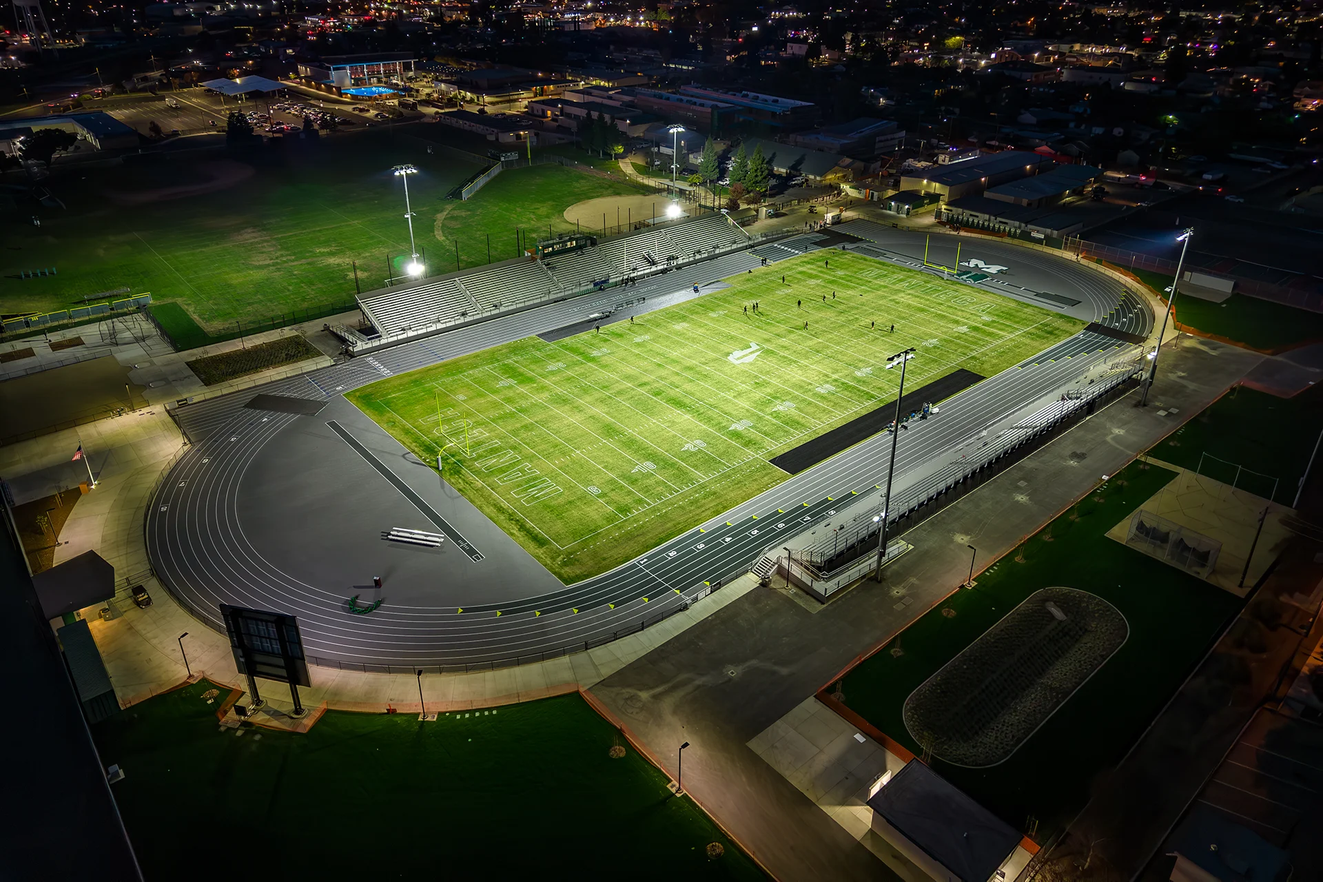 Aerial nighttime view of Manteca High School’s football stadium, running track, bleachers, and athletic facilities illuminated under stadium lights.