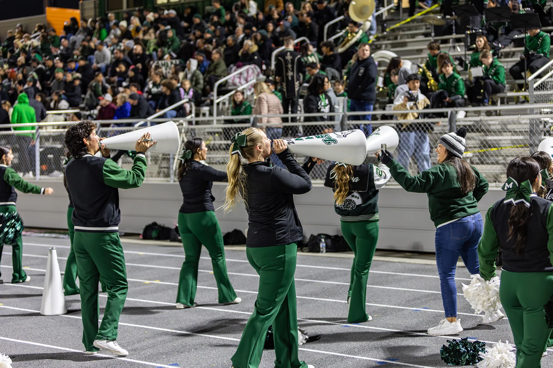 Manteca High School cheerleaders lead crowd chants from the running track during a nighttime football game with packed stadium seating in the background.