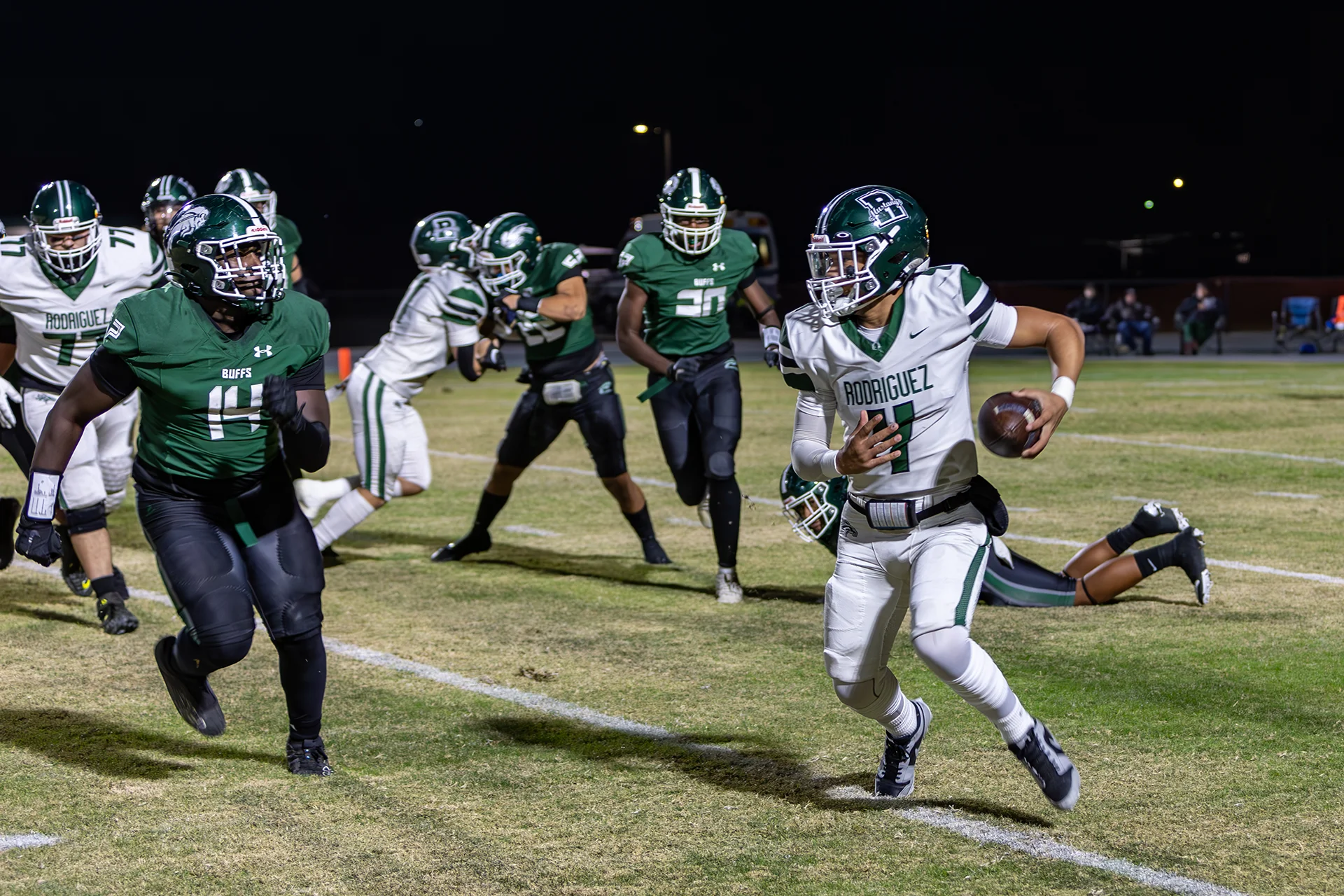 Football players compete during a nighttime game at Manteca High School as a ball carrier runs down the field under stadium lights.