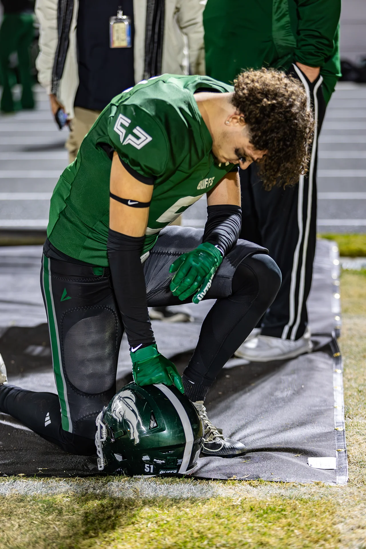 Manteca High School football player kneels beside their helmet during a pregame moment on the sideline under stadium lights.