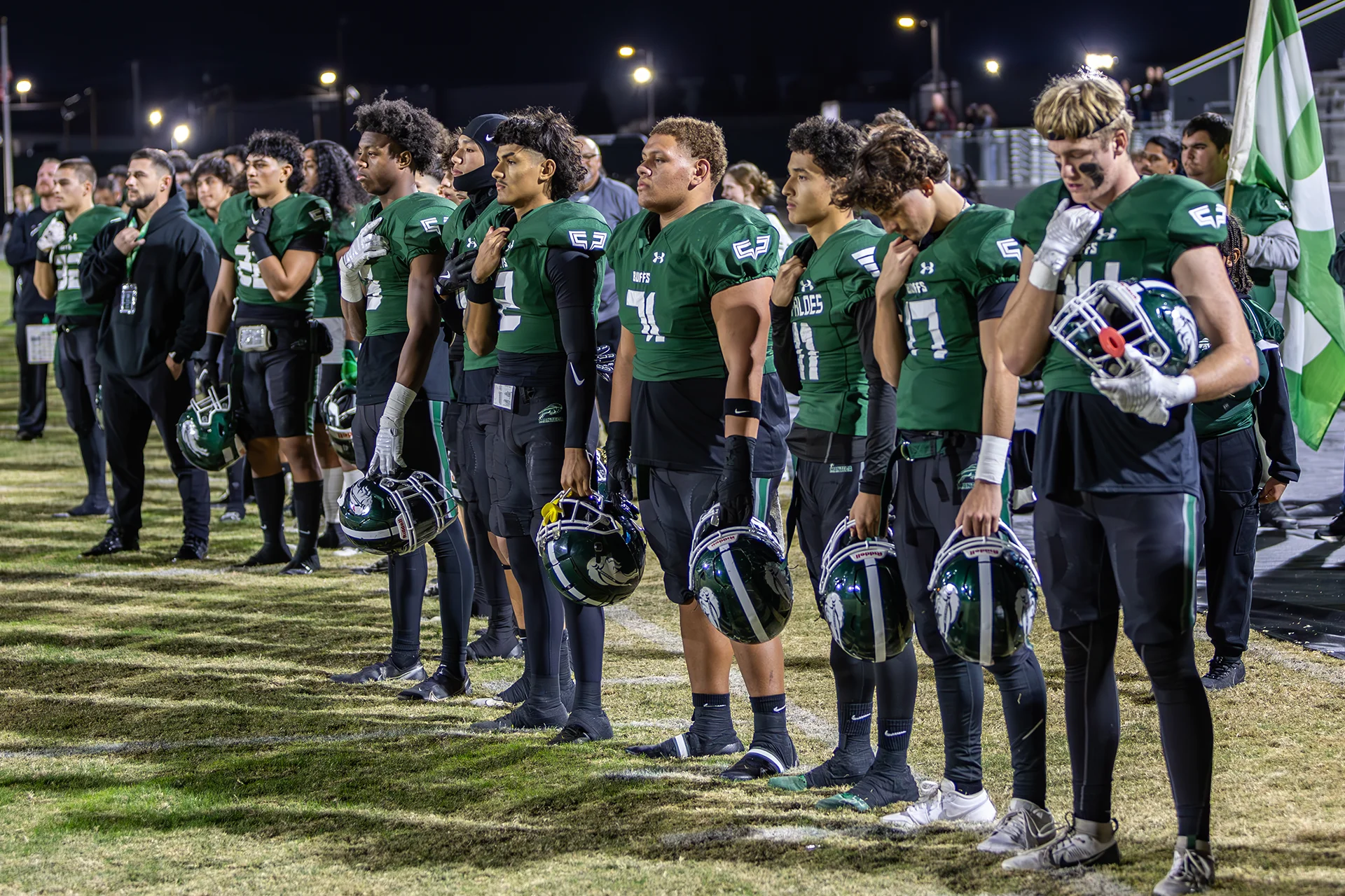 Manteca High School football players stand along the sideline during a pregame ceremony under the stadium lights.