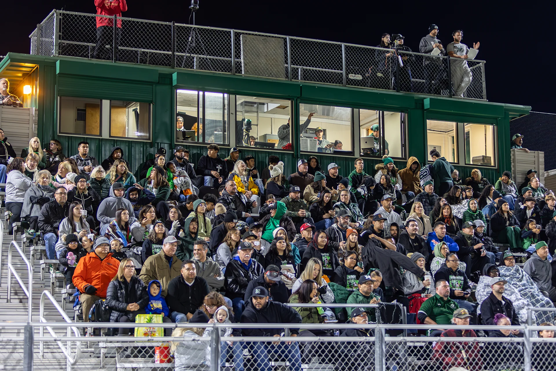 Fans fill the stadium bleachers at Manteca High School during a nighttime football game with the press box visible above the crowd.