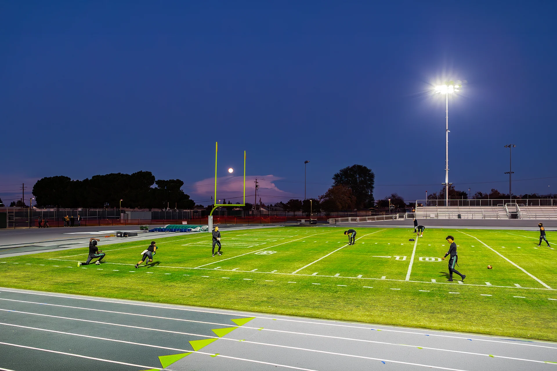 Athletes practice on the illuminated football field at Manteca High School during the evening with the track and stadium visible in the background.