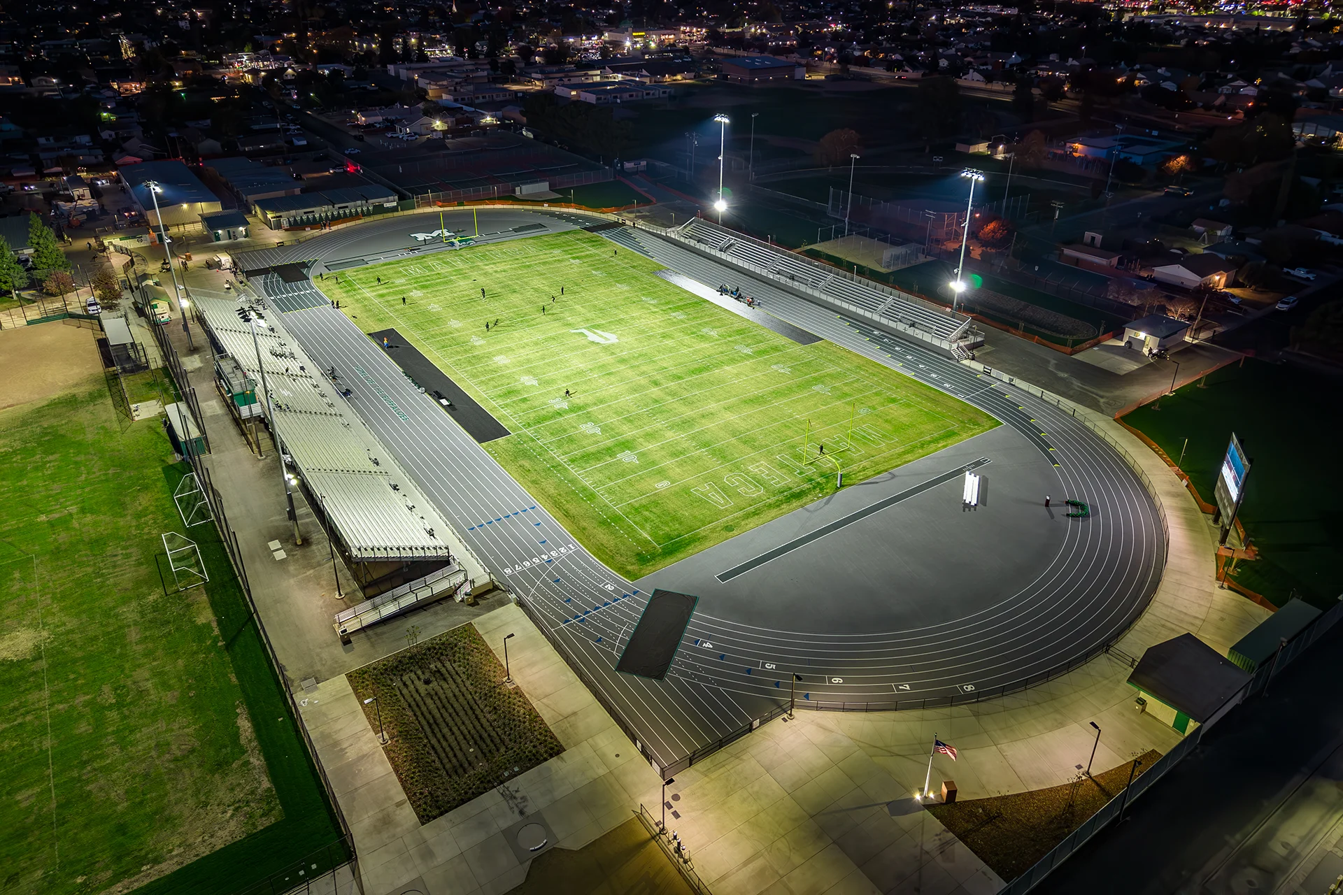 Aerial nighttime view of Manteca High School’s illuminated football stadium, running track, and athletic complex under stadium lights.