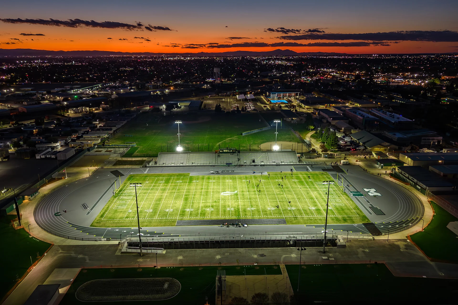Aerial view of Manteca High School’s illuminated football stadium and running track at sunset with city lights glowing in the distance.