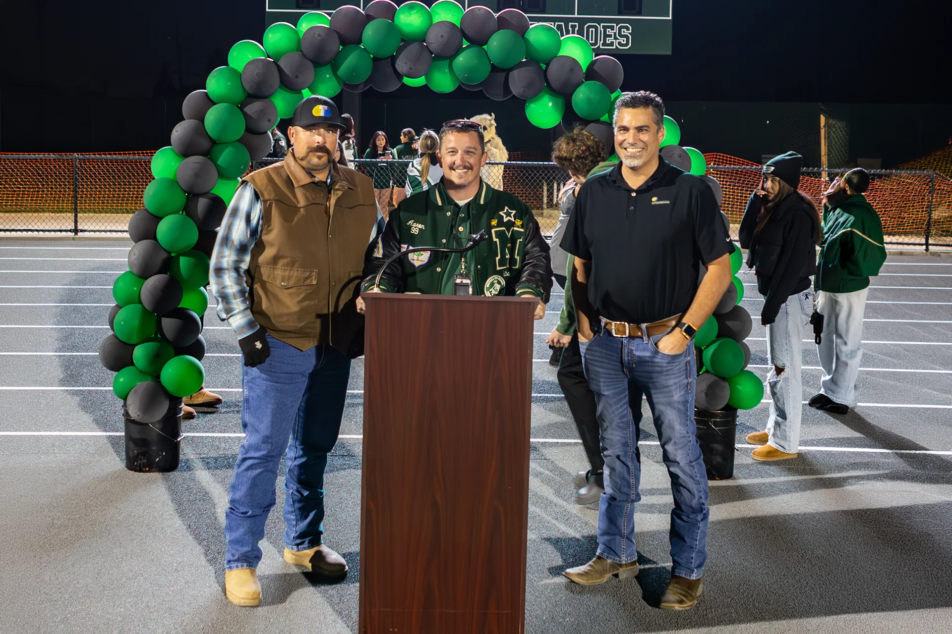 Community members stand at a podium beneath a green and black balloon arch during the Manteca High School track renovation celebration.