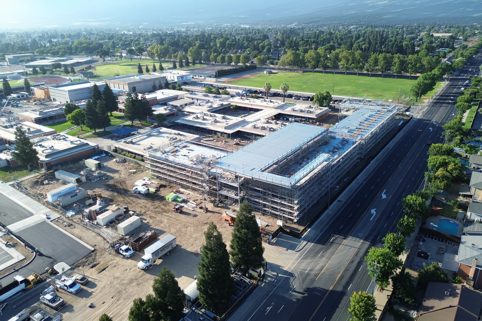 Bird's-eye drone perspective of a massive school campus construction modernization project in Manteca, California, showcasing multi-story structural steel buildings enveloped in metal piping scaffolding alongside active grading yards, supply staging areas, and concrete foundations adjacent to a main roadway.