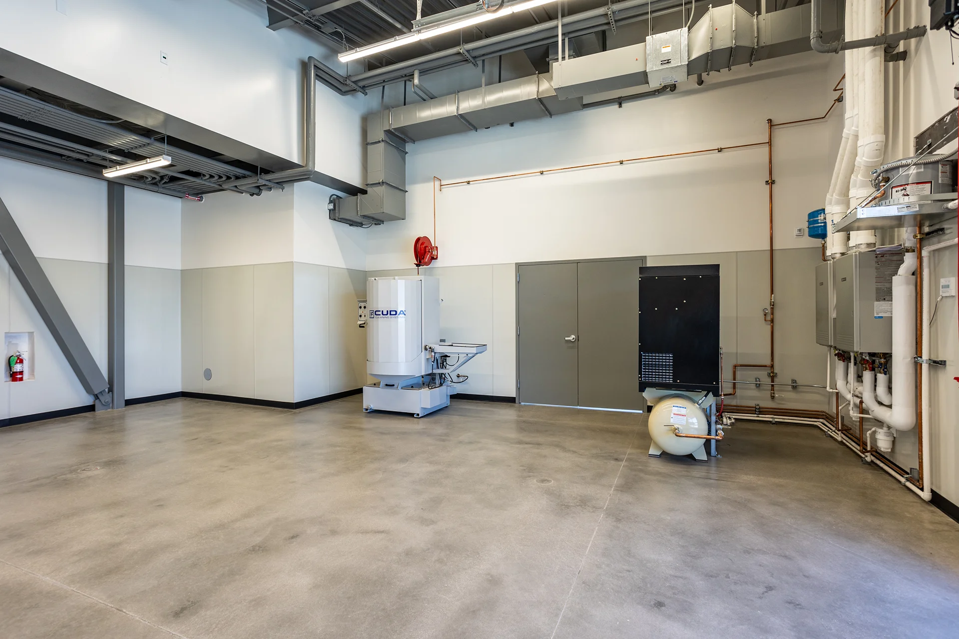 Spacious industrial training bay showcasing an array of wall-mounted commercial tankless water heaters, intricate copper plumbing manifolds, overhead galvanized HVAC ductwork, and an industrial parts washer positioned on finished concrete.