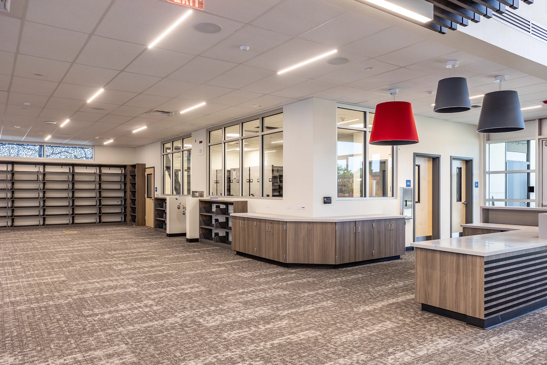 High school library reception area highlighting a wood slat and solid surface circulation counter, interior office glass windows, acoustic tile drop ceiling, and modern hanging cylinder light fixtures in charcoal gray and red.
