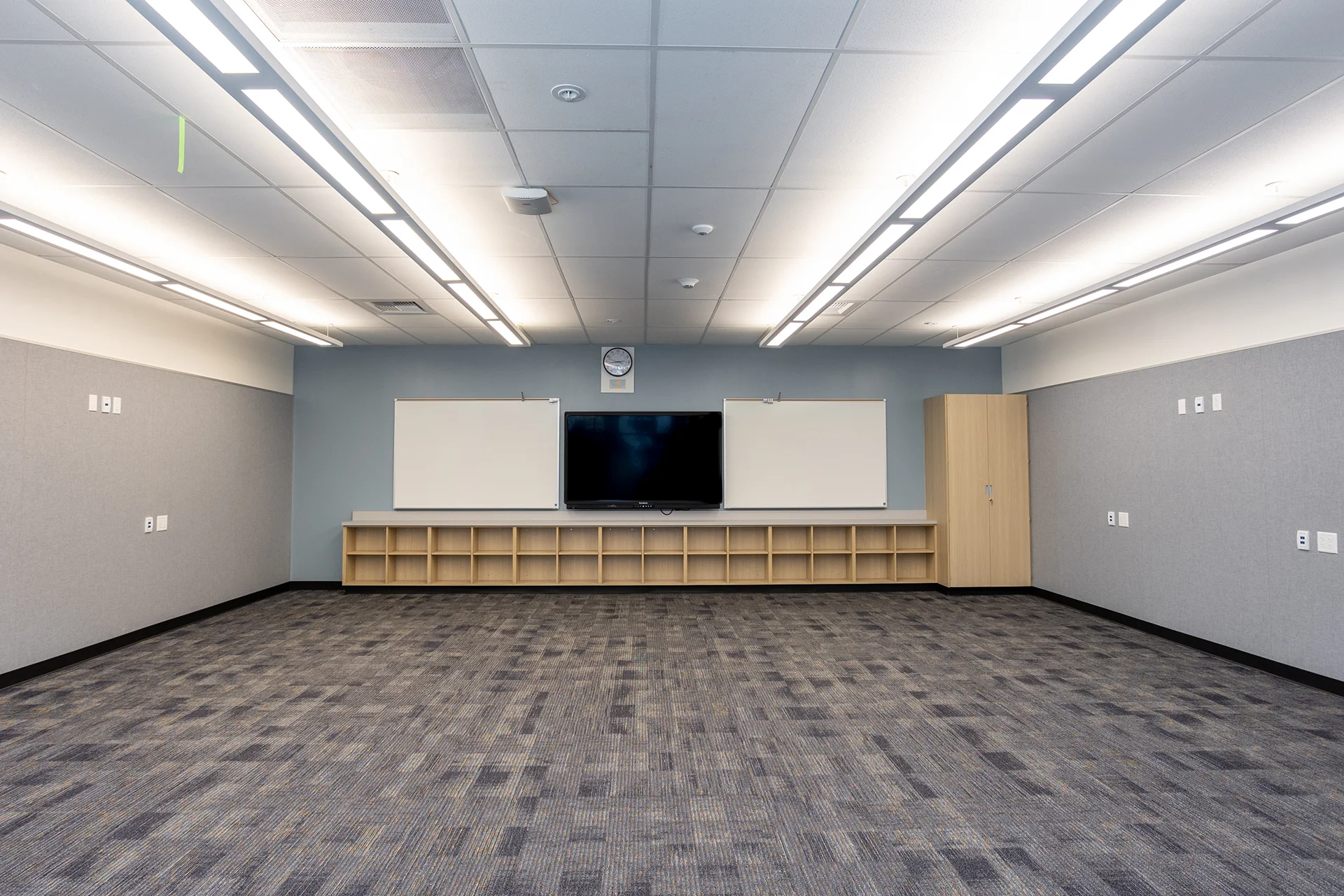 Full view of an educational lecture space displaying gray acoustic fabric wall wrap panels, a central flat-panel digital display, a light-oak base storage credenza with matching tall storage tower, and commercial carpet tile flooring under flush-mount LED troffers.