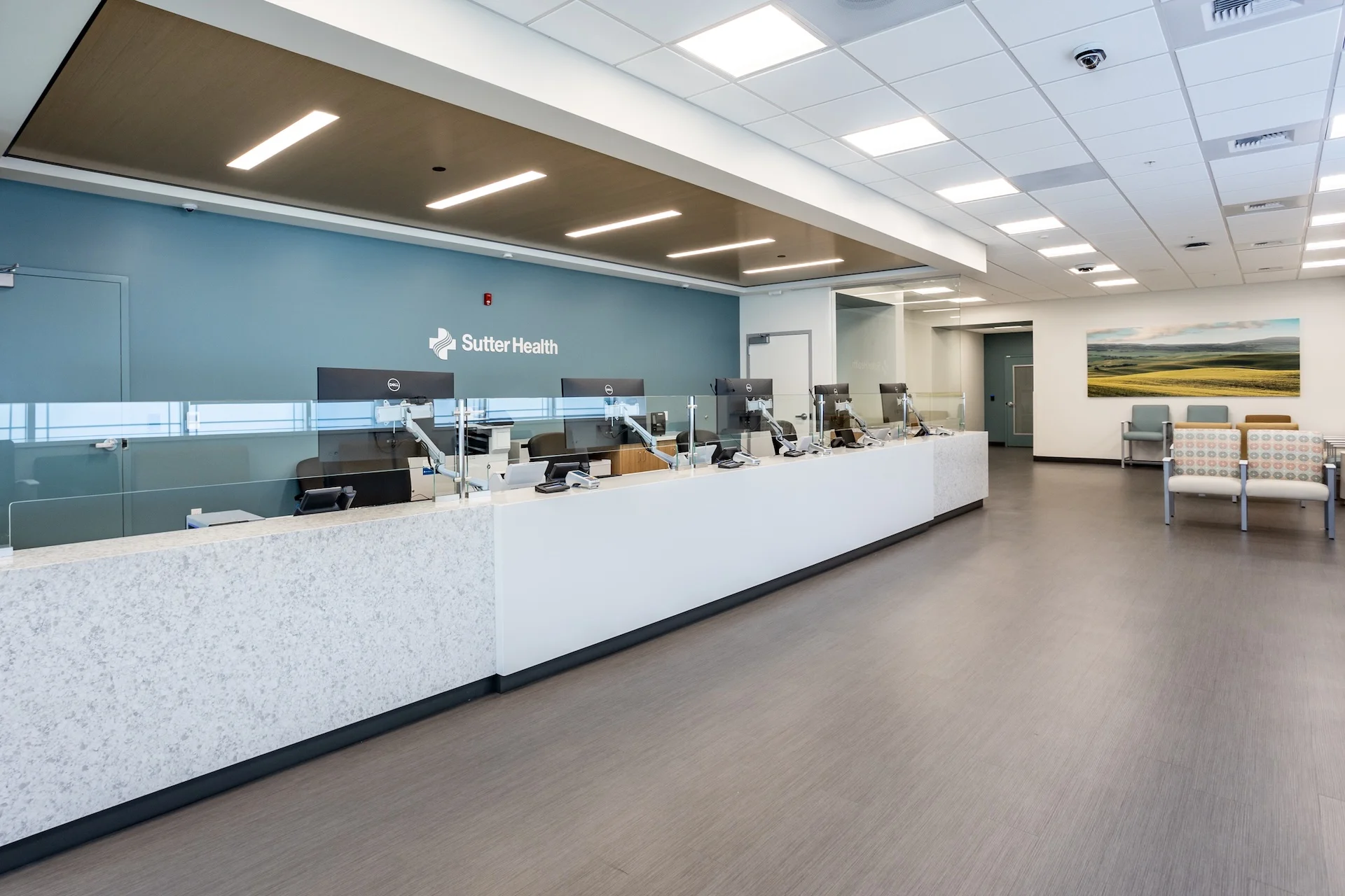 Wide-angle interior photograph showcasing a large, modern healthcare reception desk in Lathrop, California, featuring polished grey solid surface countertops, light wood veneer soffit with linear LED lighting, several integrated computer workstations with monitors, and glass privacy dividers, looking toward a patient waiting area with soft seating.