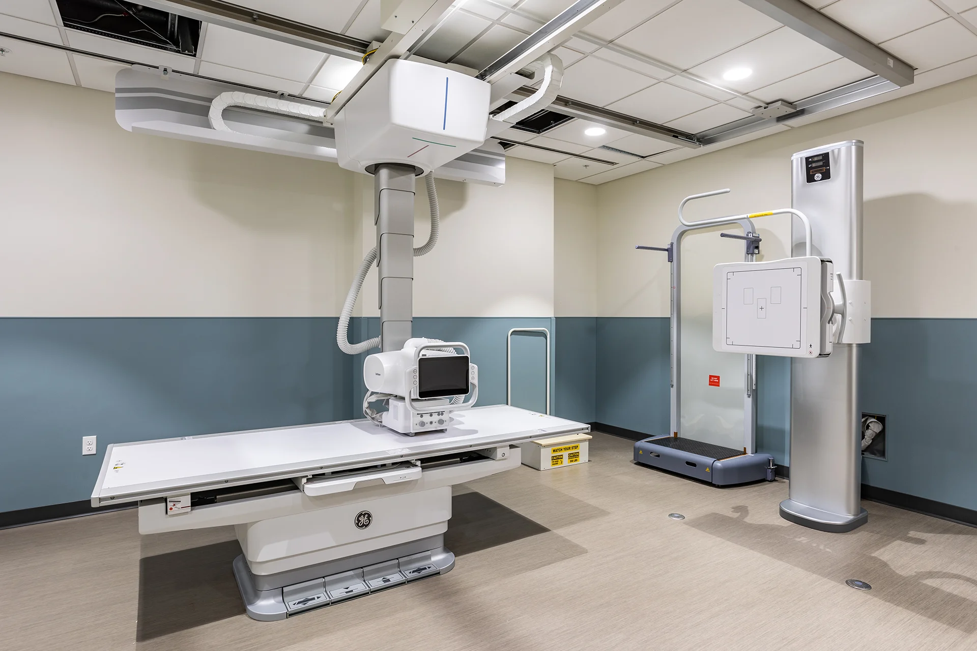 Professional radiography room featuring a ceiling-mounted GE digital X-ray machine with an adjustable patient table, a wall-mounted chest bucky stand, a patient safety platform with handrails, and protective teal wall paneling.