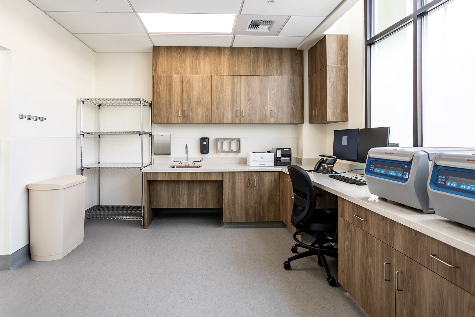 Healthcare laboratory workspace featuring an L-shaped solid-surface countertop, an ADA-compliant cutout sink, built-in walnut-finished upper and base cabinetry, desktop computers, Thermo Scientific benchtop centrifuges, and seamless commercial vinyl flooring.