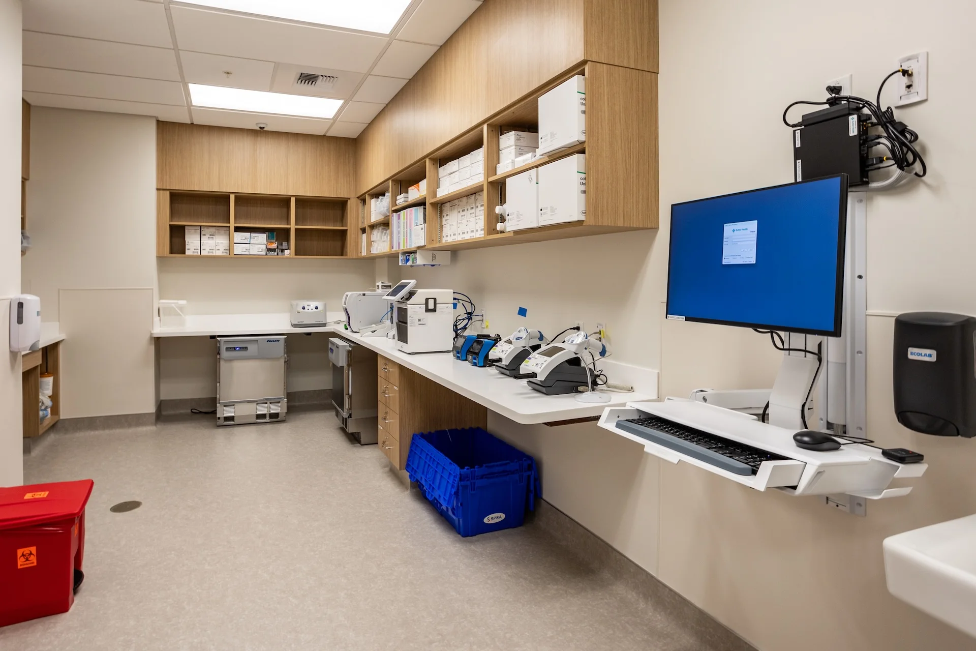 Inside a clinical medical lab space featuring custom wood grain cabinetry, seamless solid surface countertops, white commercial grade sterilizers and centrifuges, a computer charting station with an ergonomic keyboard tray, and blue medical supply bins on the resilient floor.