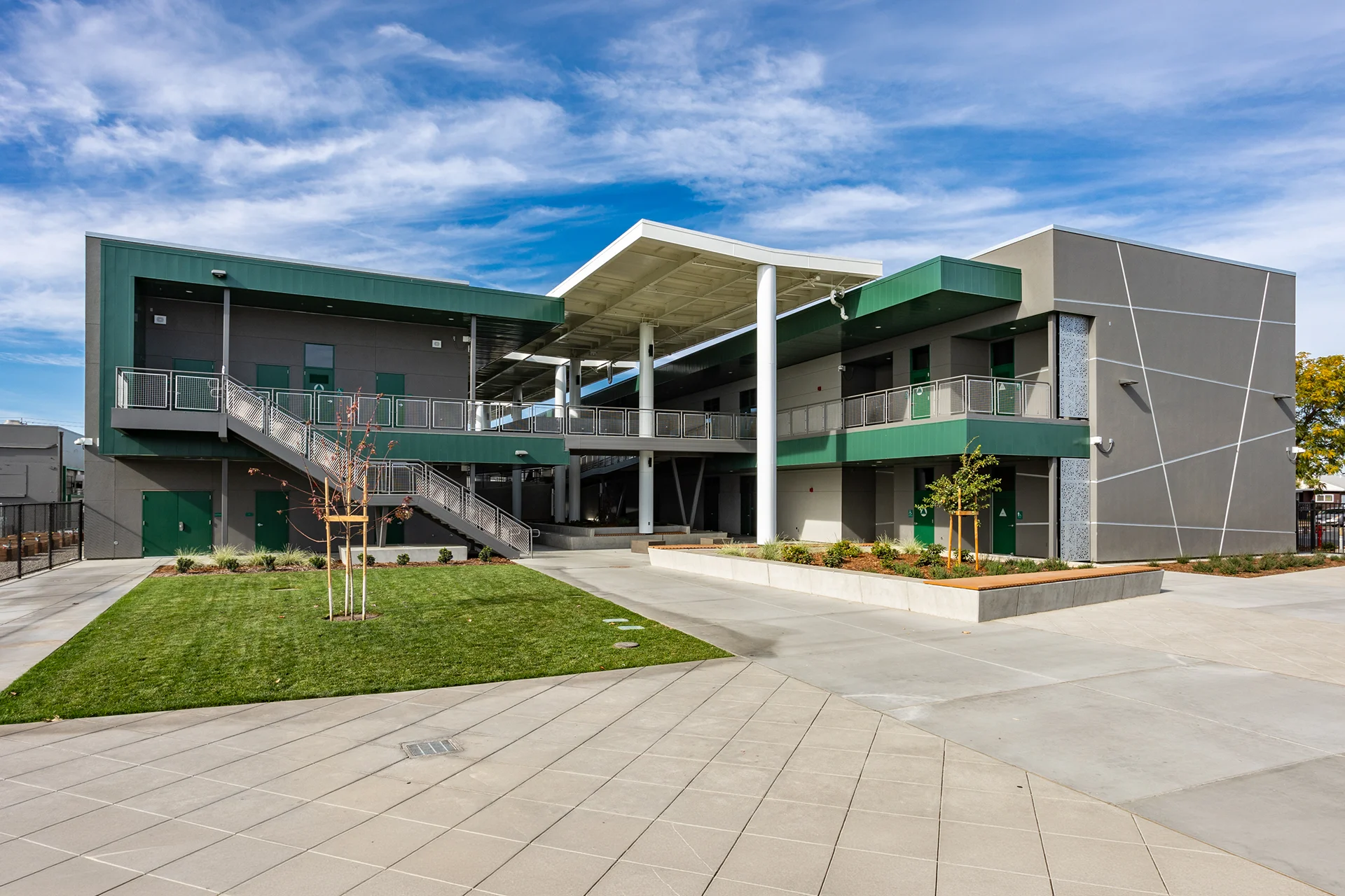 Eye-level view of a newly developed campus quadrangle in San Joaquin County, featuring decorative geometric concrete paving, built-in concrete planters, a central lawn, open-air multi-level walkways, and accent walls with geometric white detailing.