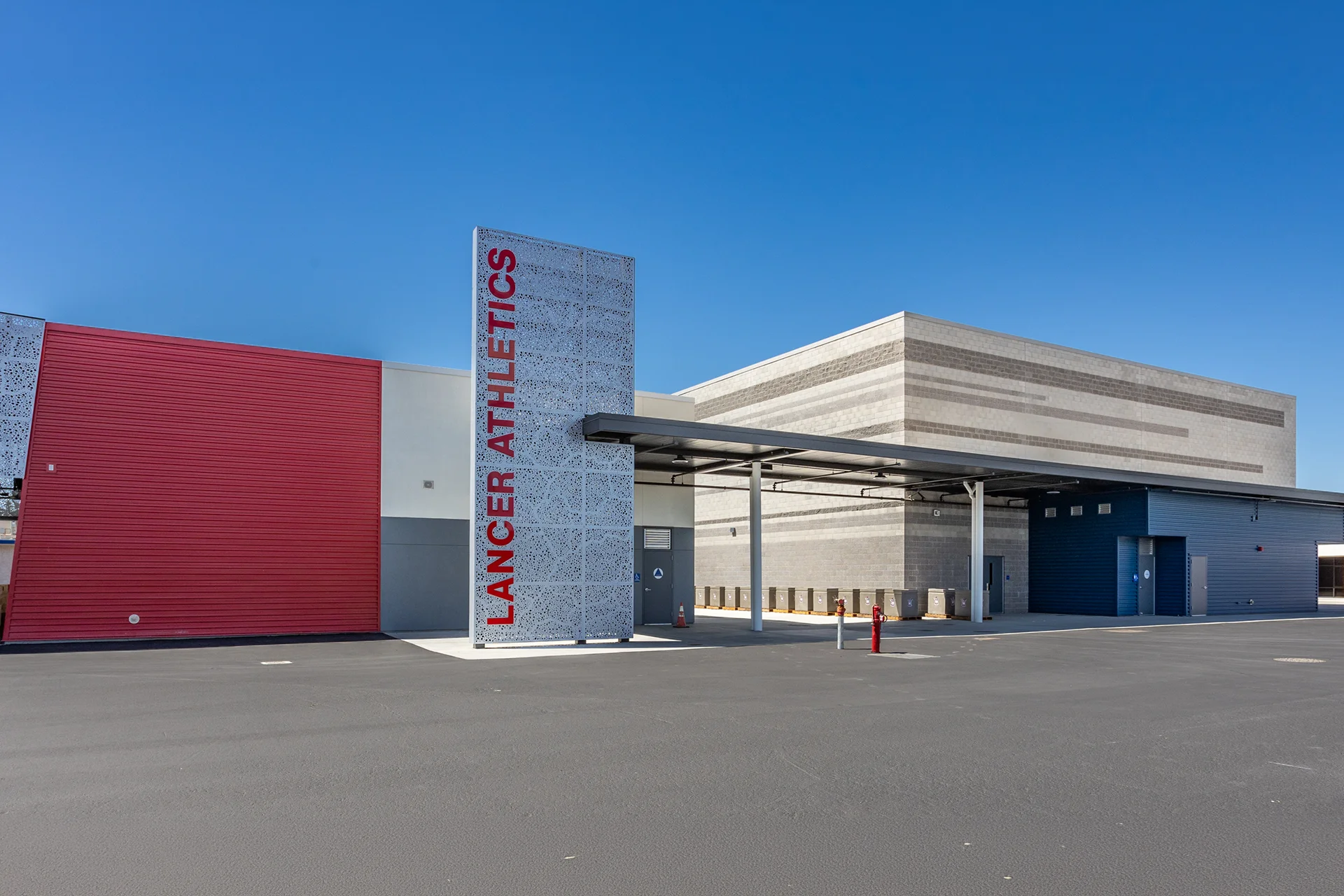 Commercial athletic facility exterior in Manteca, California featuring a perforated metal monument sign tower, red corrugated steel siding, a covered concrete walkway, and split-face concrete masonry unit walls.