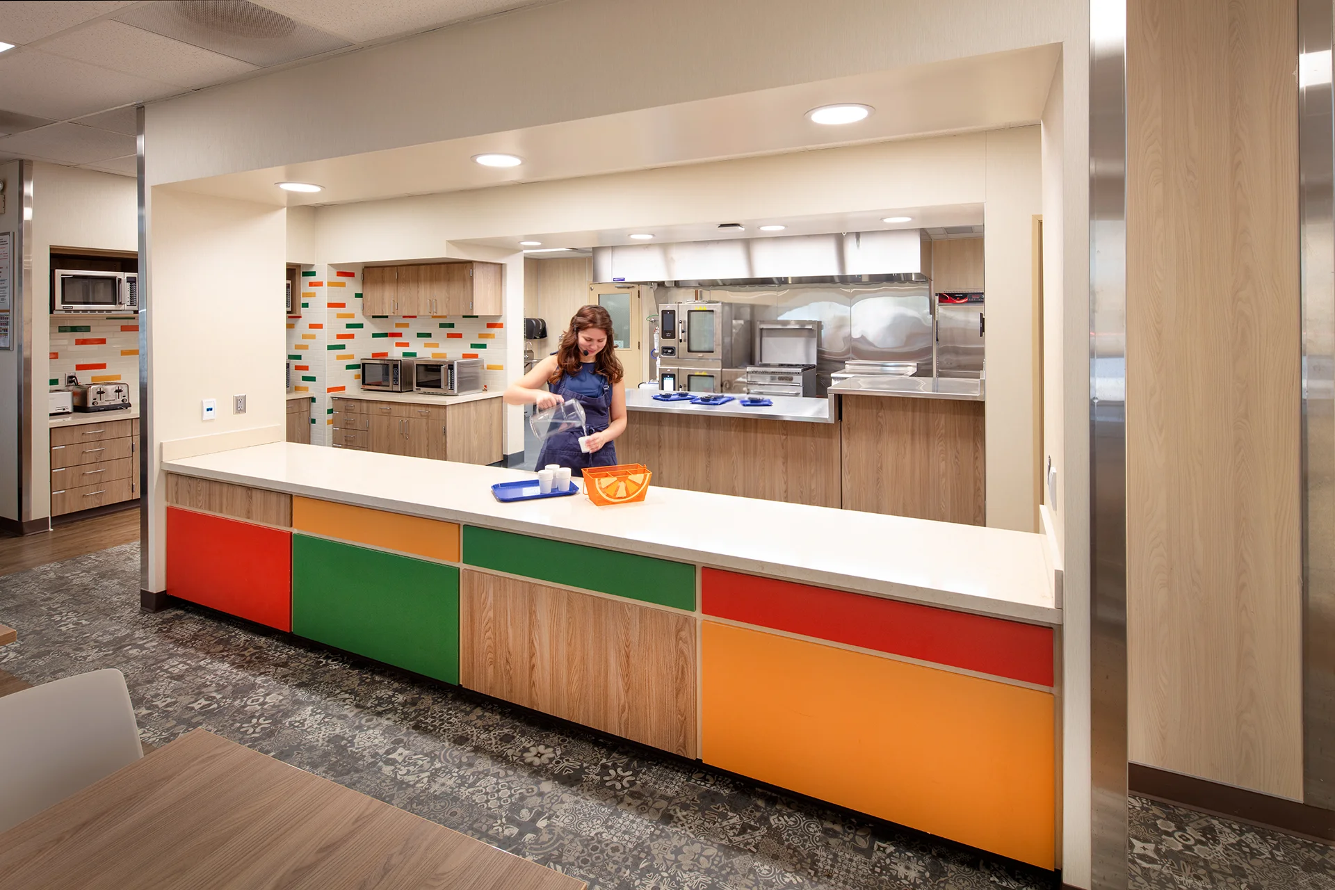 Commercial food service counter installation featuring custom wood laminate cabinetry, solid surface white countertops, vibrant multi colored accent panels, and a stainless steel prep kitchen visible in the background.