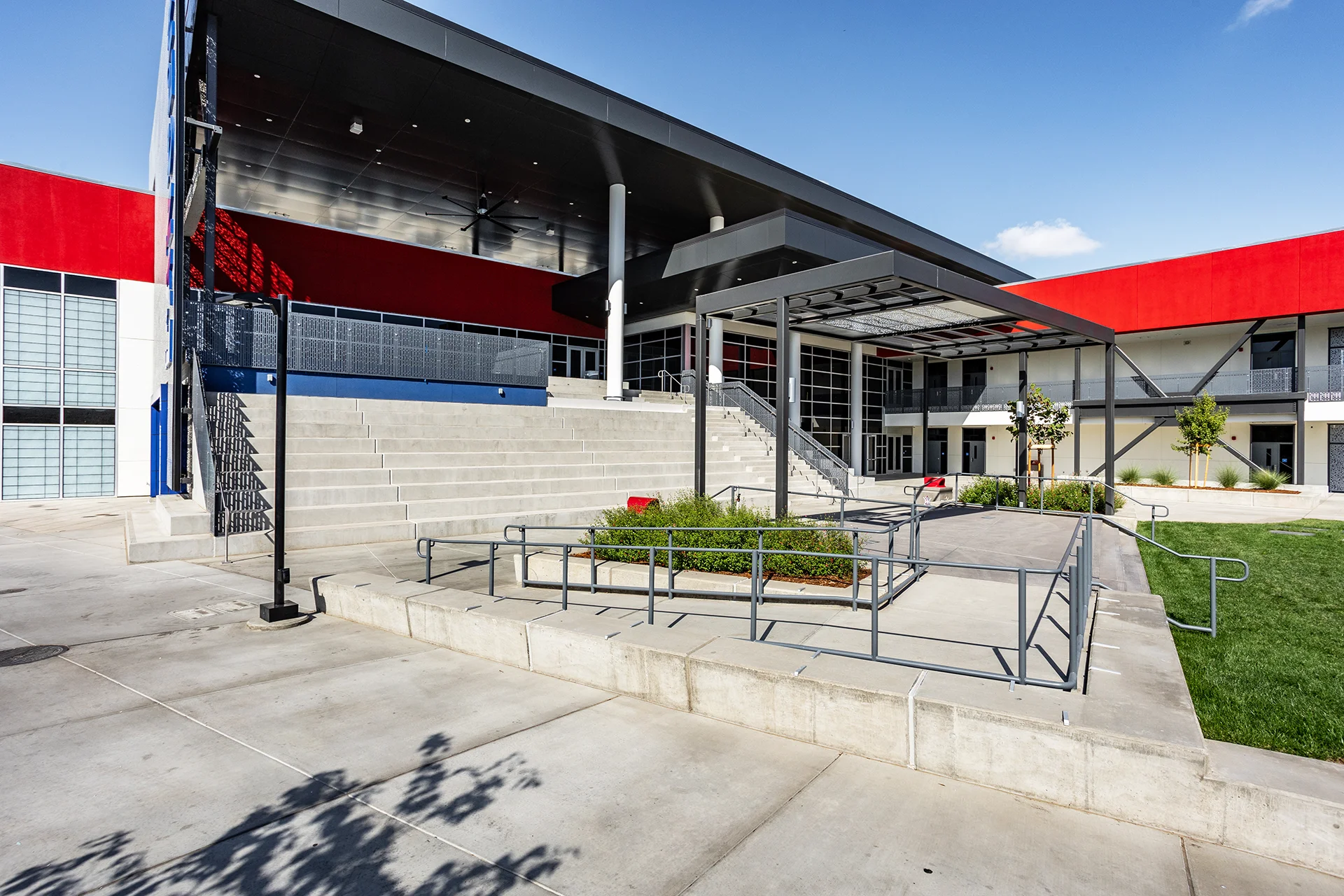 Ground-level view of an institutional concrete stadium staircase assembly flanked by a multi-line grey metal handrail system and a low retaining wall layout. The background highlights an expansive dark metal roof overhang canopy with large industrial ceiling fans and white column pillars.