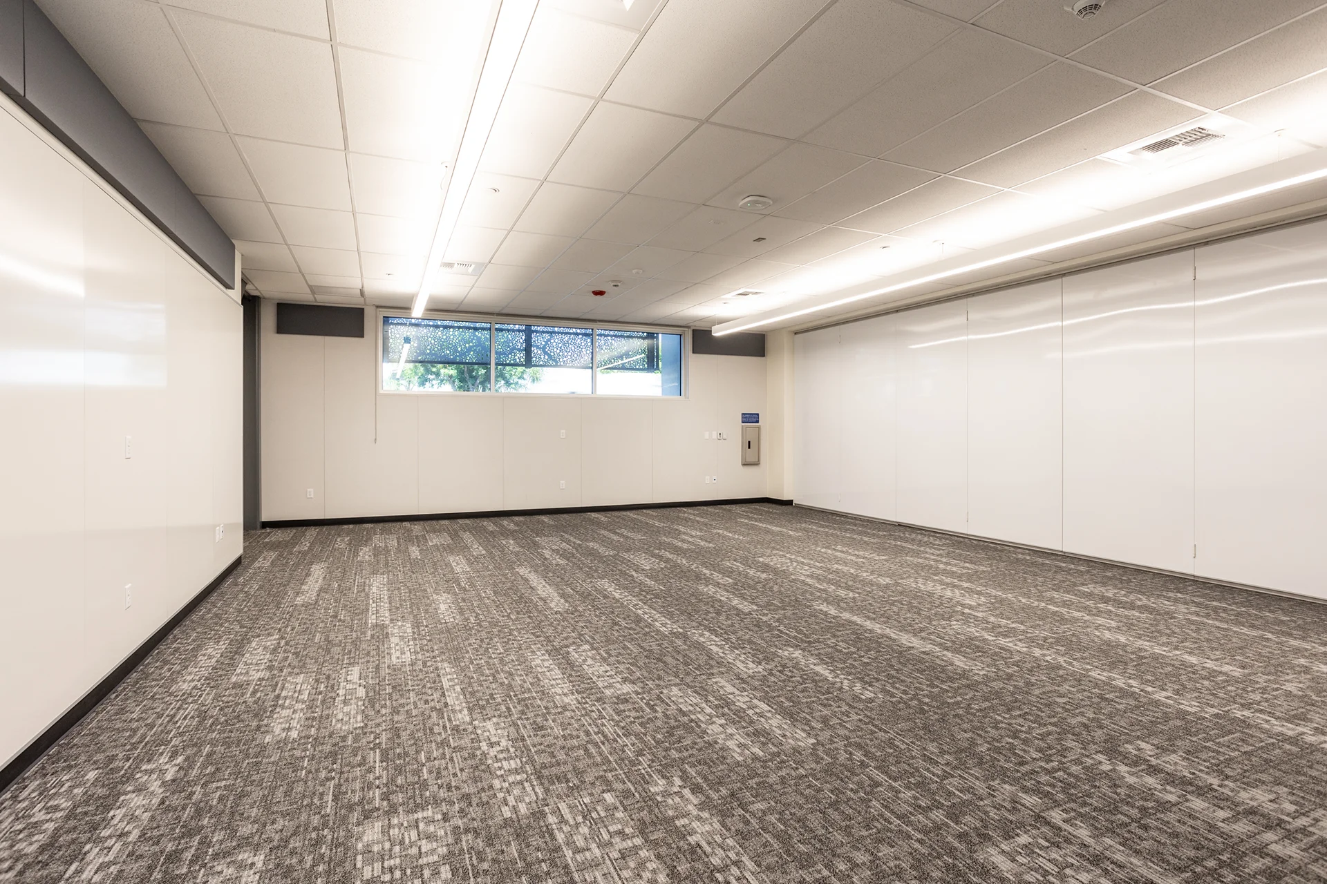 Commercial high school classroom renovation featuring expansive floor-to-ceiling dry erase whiteboard walls, gray linear carpet tiles, an acoustic drop ceiling with strip LED lighting, and a wide perimeter window.