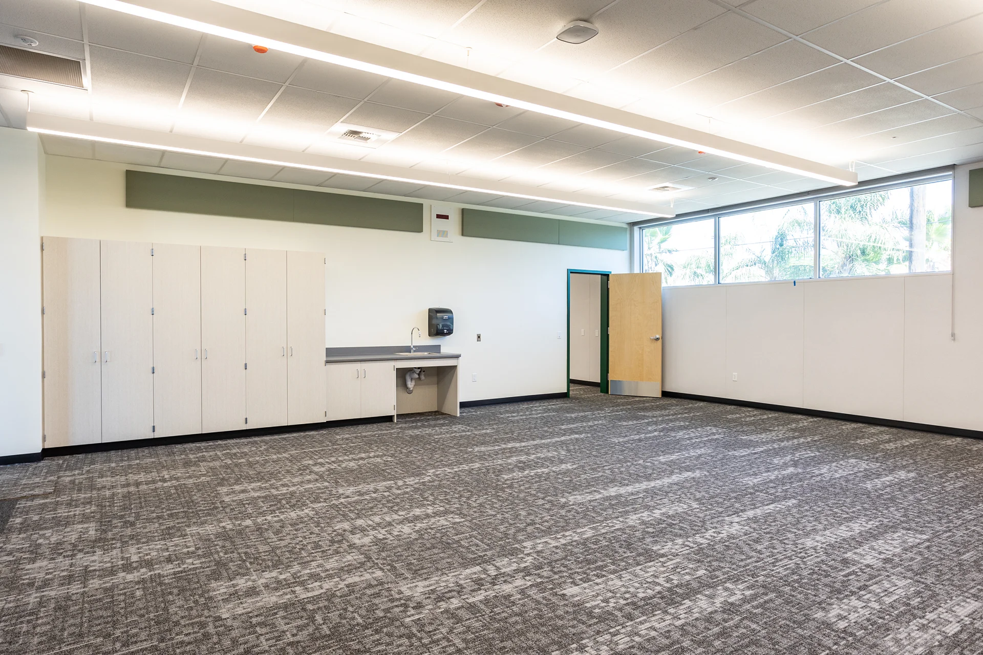 Interior perspective of a flexible classroom space in Manteca, California, displaying built-in light maple laminate storage lockers, an ADA compliant recess sink counter, high clerestory safety windows, and heavy duty commercial grade carpeting.