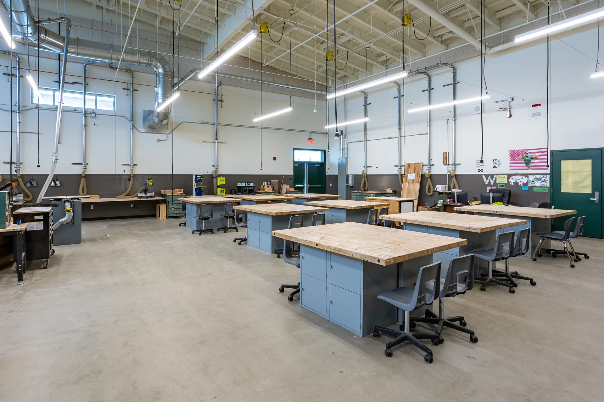 Interior view of a newly constructed institutional woodshop classroom, featuring rows of heavy-duty butcher block workbenches over steel storage bases, rolling office chairs, overhead dust collection ductwork, exposed ceiling joists with pendant LED lighting, and green metal exterior exit doors.