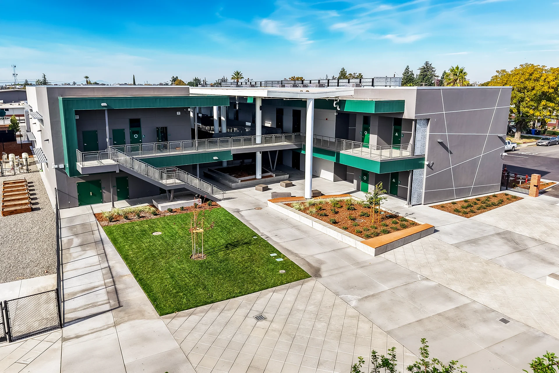 Expansive drone view of an educational plaza featuring modern two-story academic blocks, an open canopy roof covering an interior courtyard, decorative concrete paving, multiple green commercial steel exit doors, and an accent wall featuring sharp white geometric reveal lines.