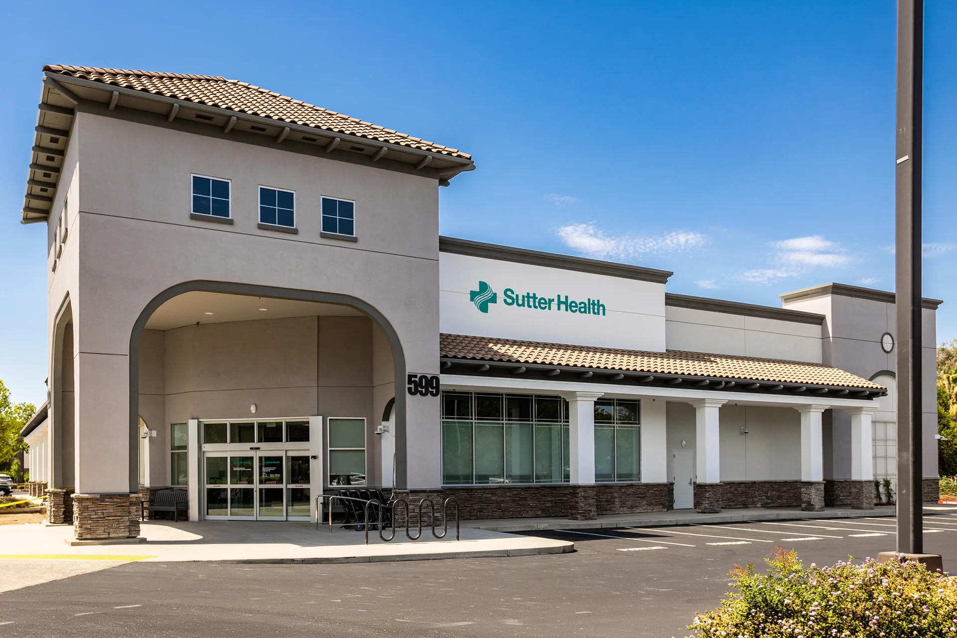 Wide exterior shot of a commercial healthcare facility building in Lathrop, California, showcasing architectural stucco elements, concrete columns with stacked stone bases, a clay tile roof awning, a prominent arched driveway canopy, and clear storefront glass windows under a bright blue sky.