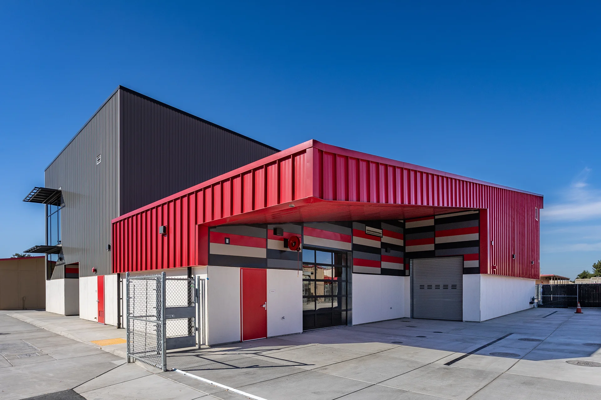 Exterior corner view of a contemporary commercial building featuring dual-tone corrugated metal siding, a prominent bold red cantilevered roof canopy, a segmented glass roll-up garage door, and perimeter security chain-link fencing over concrete paving.
