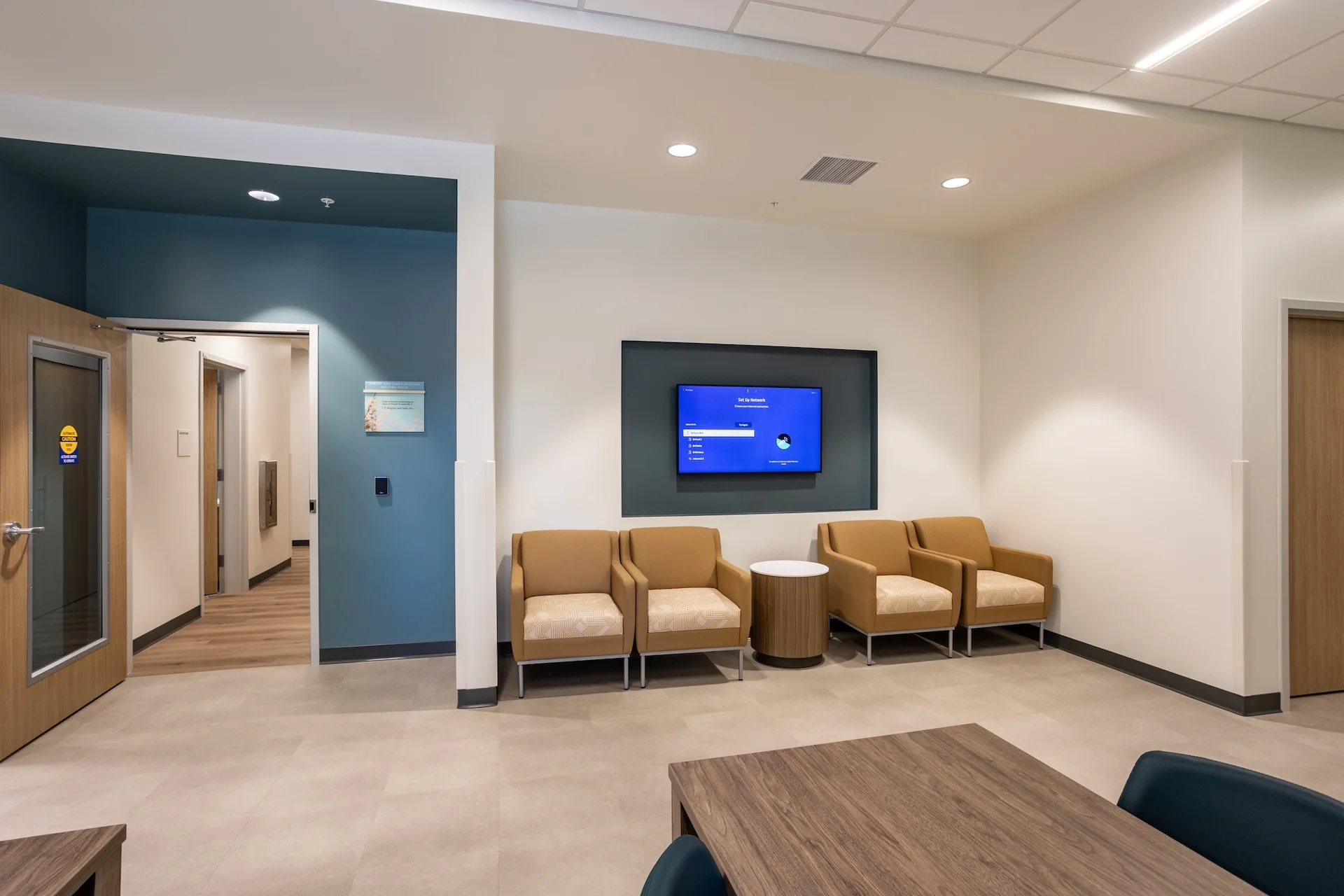 Interior view of a medical clinic waiting nook featuring tan upholstered armchairs, a wall-mounted digital display set within a recessed accent alcove, neutral acoustic ceiling tiles with recessed LED downlights, and premium vinyl tile flooring leading past an automatic sliding glass entryway door.