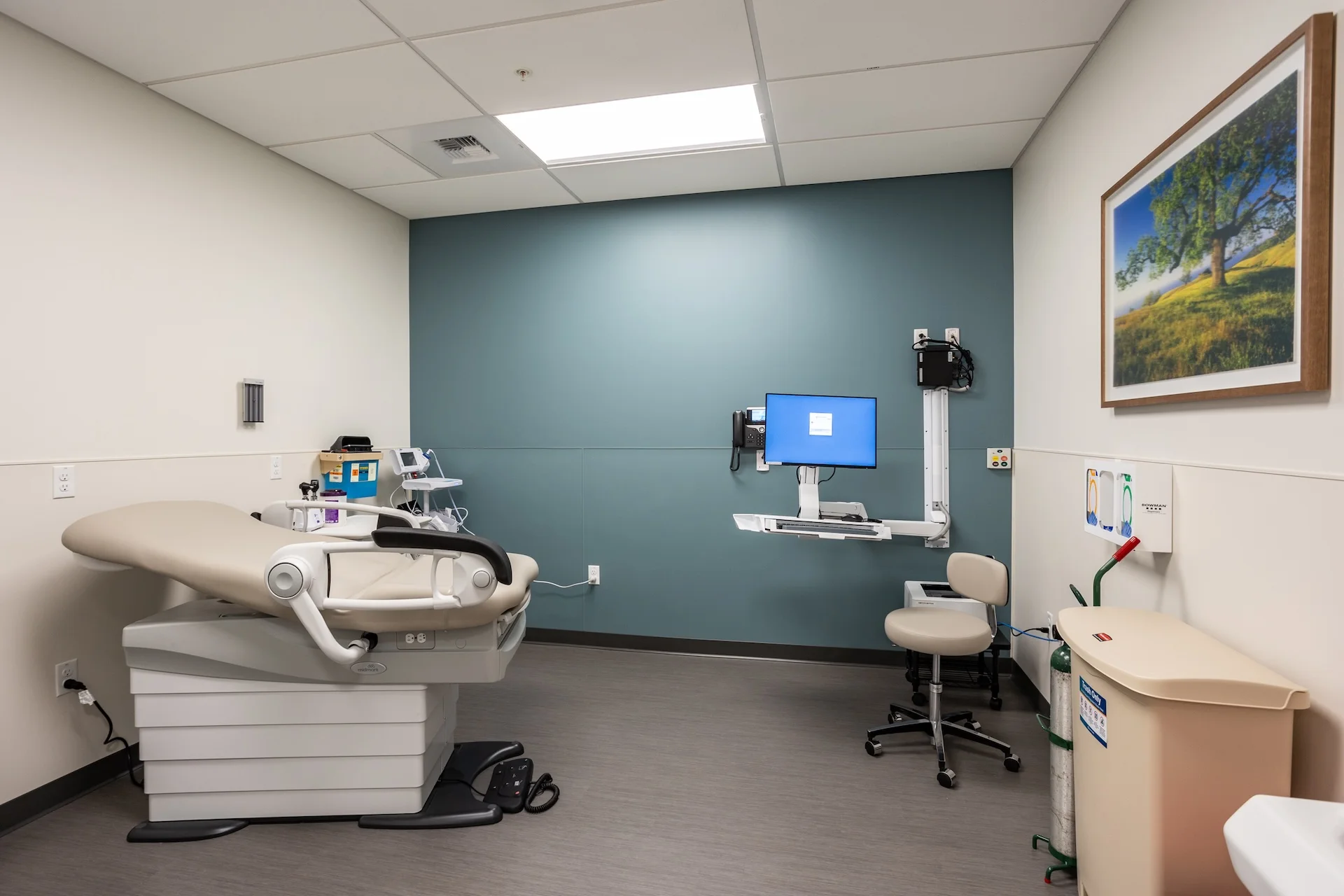 Interior of a complete medical exam room showing a motorized patient table, a physician's rolling stool, a wall-mounted computer charting station with dual monitors, a diagnostic otoscope/ophthalmoscope set, integrated sinks, a framed landscape photograph, and turquoise accent walls.