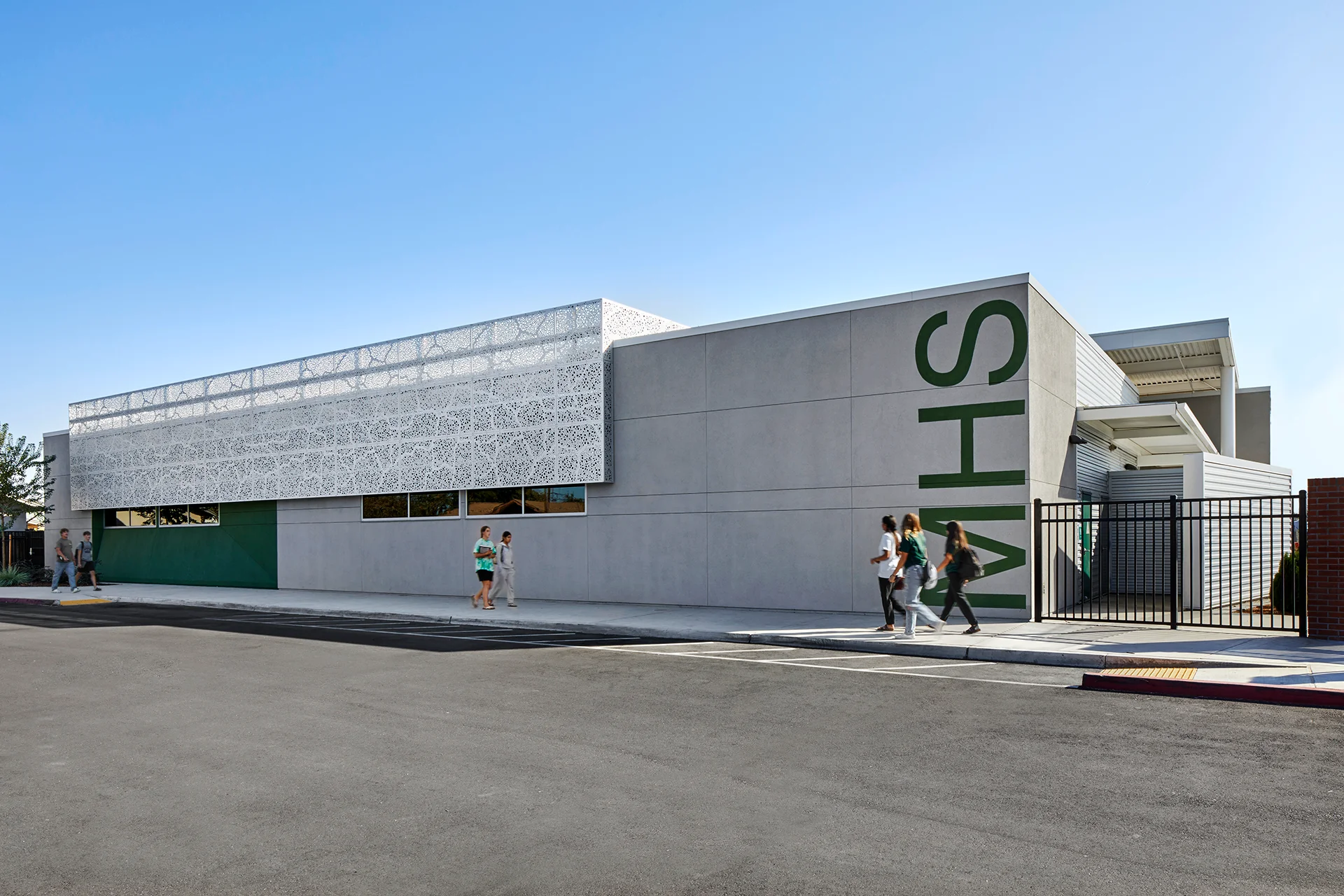 Side profile view of a contemporary school building in San Joaquin County, featuring smooth gray stucco panels accented by massive dark green inlaid block lettering graphics, an integrated line-pattern metal sunscreen panel, and a black steel security perimeter fence.