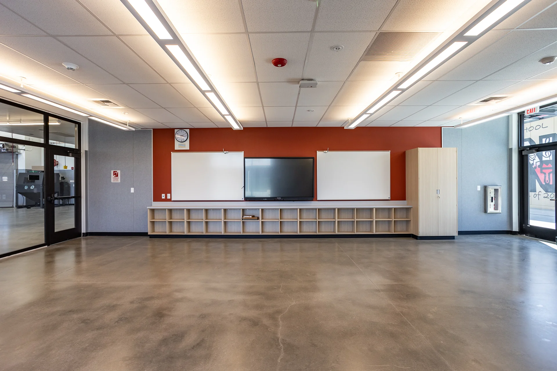 Symmetrical view of a technical training classroom featuring polished concrete flooring, a central digital smart display flanked by dry-erase whiteboards, custom light-wood base cabinetry, and a deep accent wall beneath recessed commercial LED strip lighting.