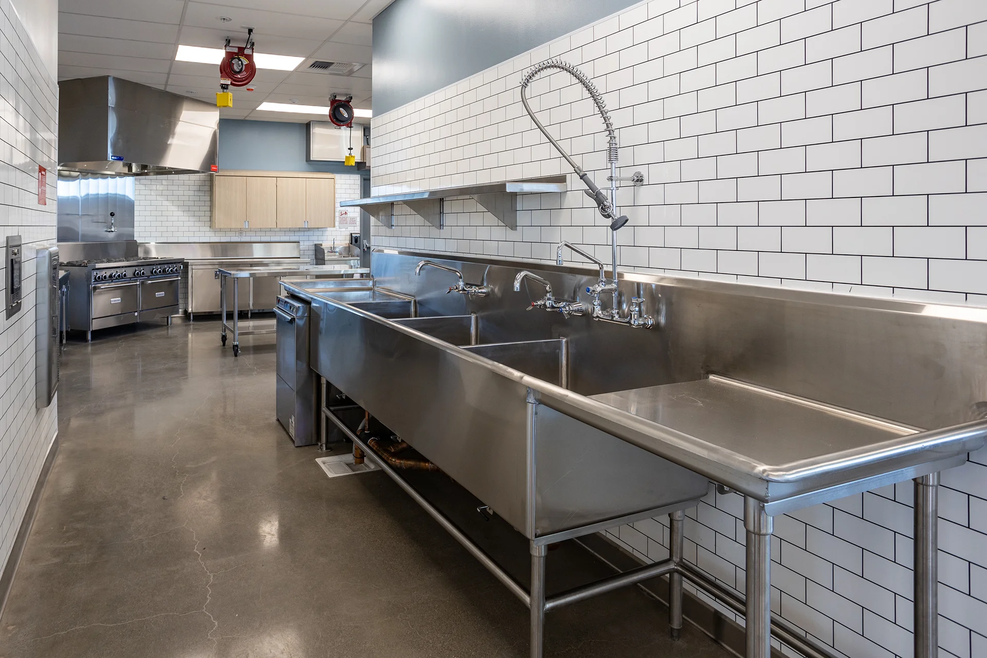 Deep three-compartment industrial wash sink featuring an overhead spring-action pre-rinse spray assembly and wall-mounted faucets, installed against a full-height white subway tile accent wall with overhead stainless steel shelving.