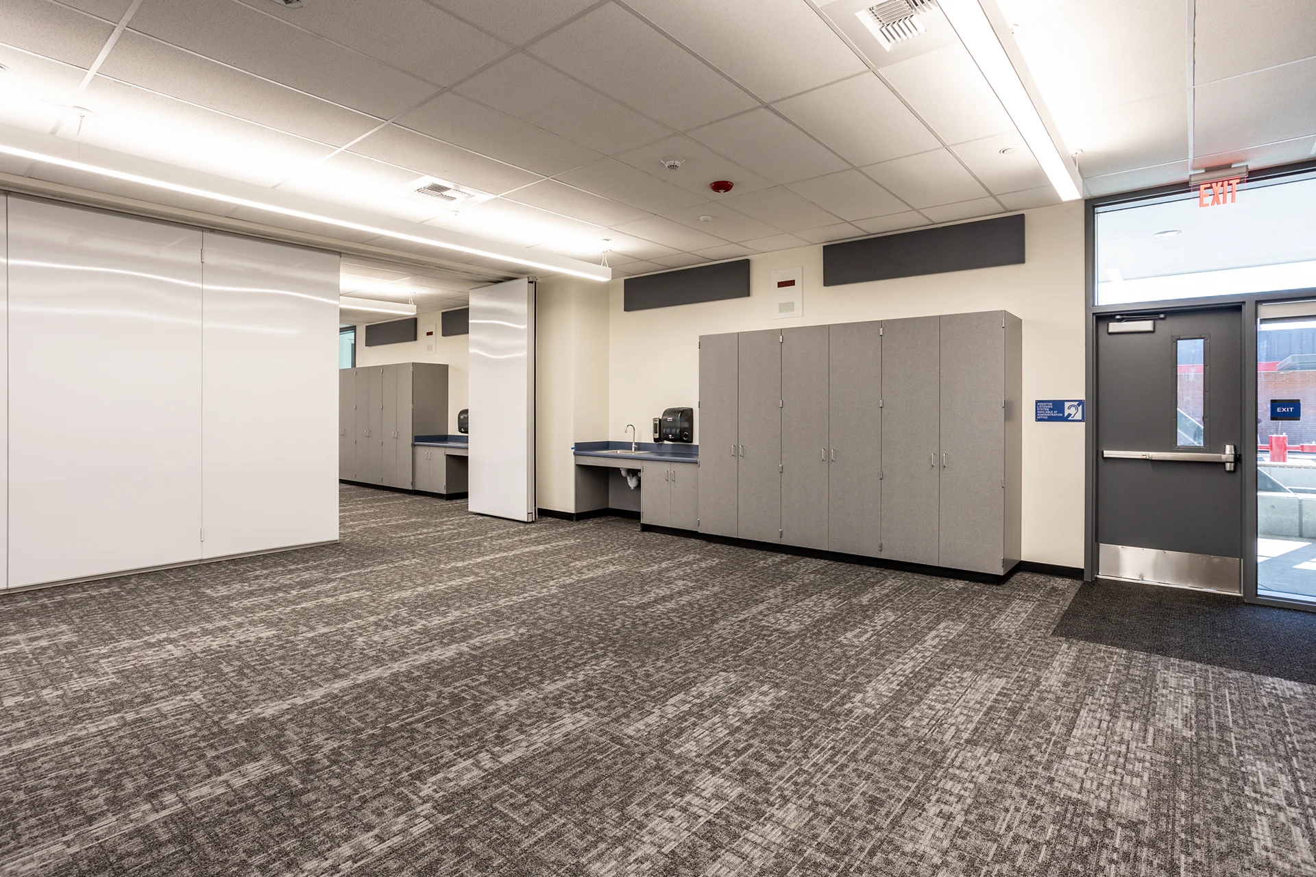 School instructional room displaying built-in gray laminate storage cabinets, a perimeter sink station, mobile whiteboards, and a steel exterior exit door under an illuminated exit sign.